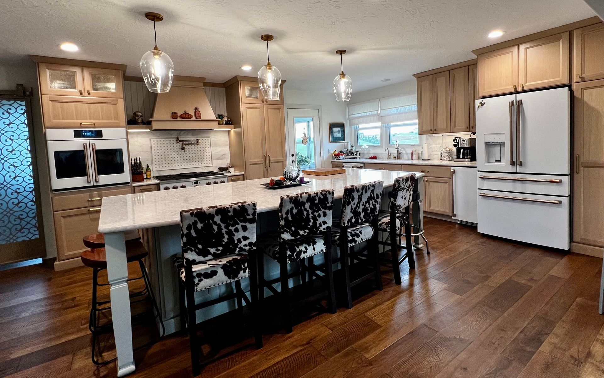 A kitchen with wooden cabinets, a large white island with cow-print stools, and a white refrigerator on wood flooring.