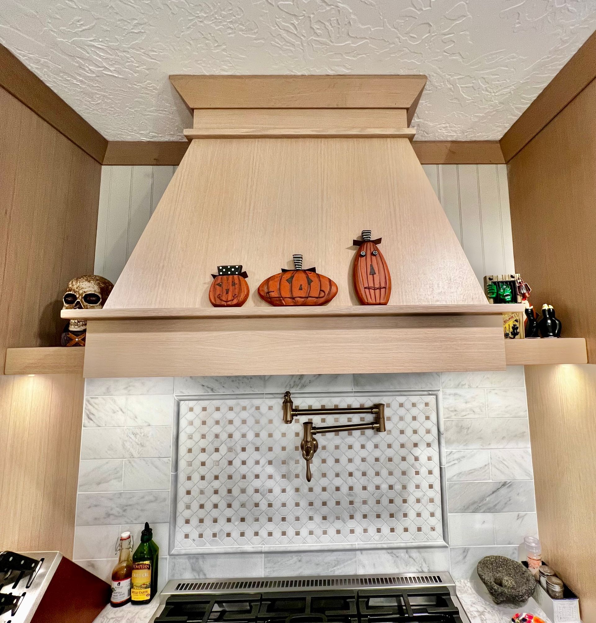Wooden kitchen range hood decorated with three orange pumpkins and a skull, over a marble tile backsplash and stove.