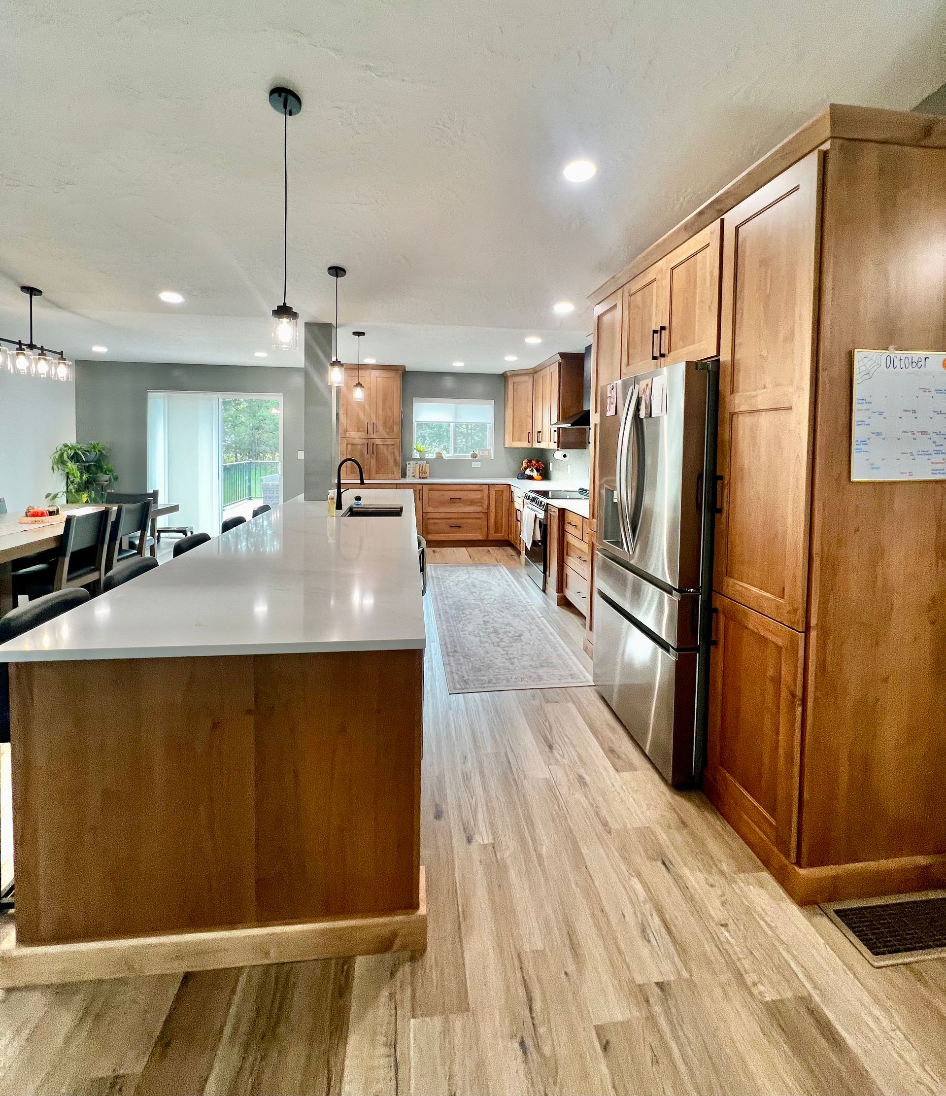 A modern kitchen with wood cabinets, a large white-topped island, stainless steel refrigerator, and wood-look flooring.