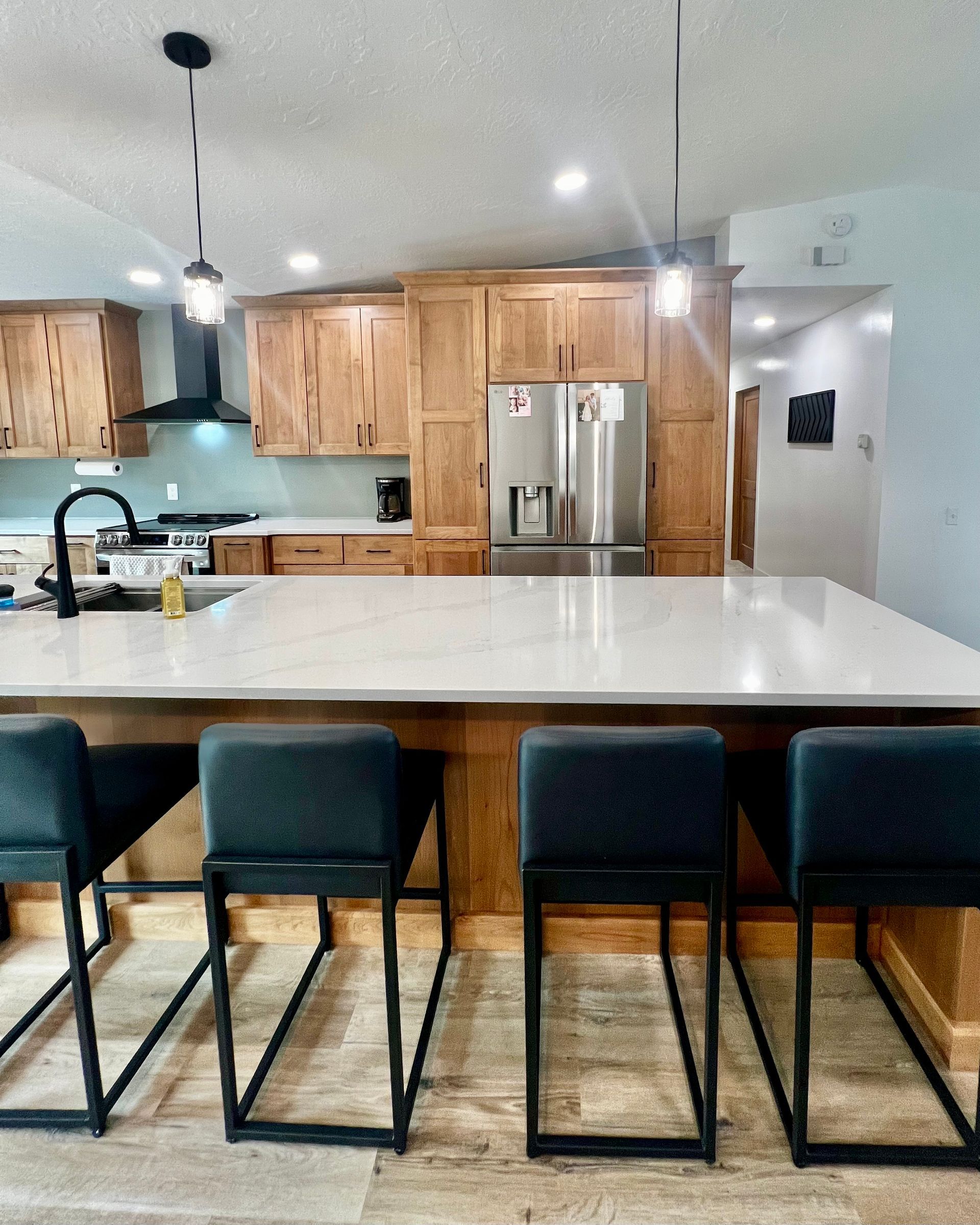 A modern kitchen features wooden cabinets, a large white countertop island, four dark stools, and two hanging lights.