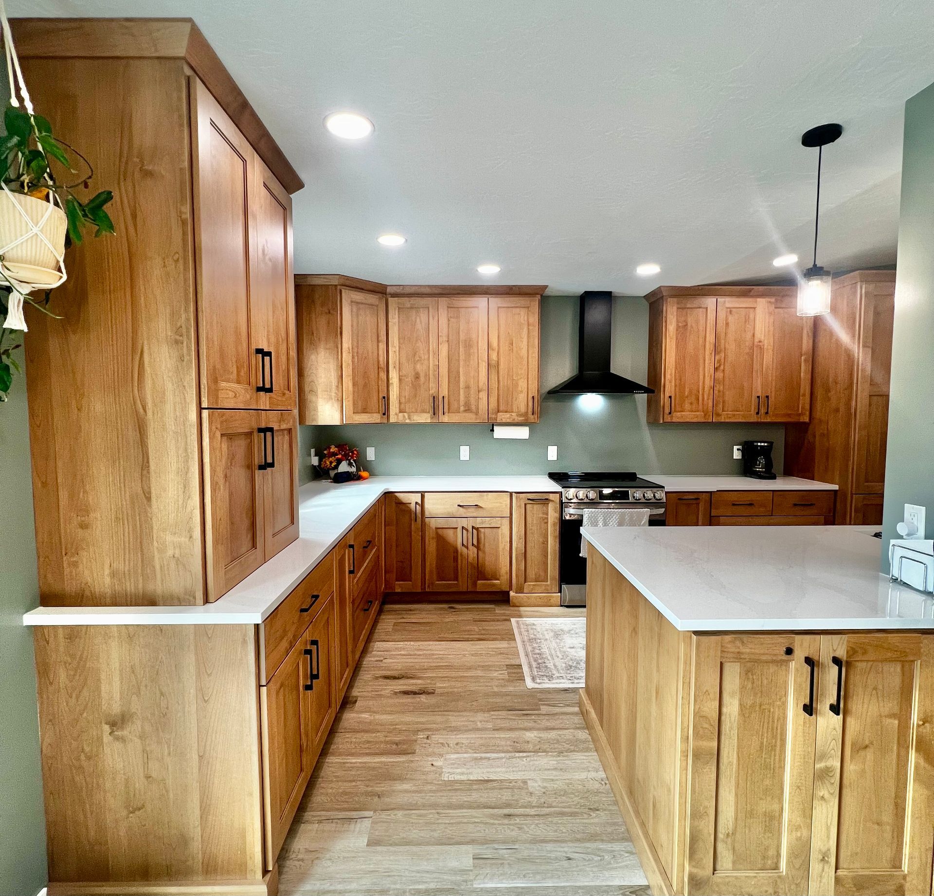 A modern kitchen with warm-toned wooden cabinets, white quartz countertops, a black range hood, and light wood flooring.