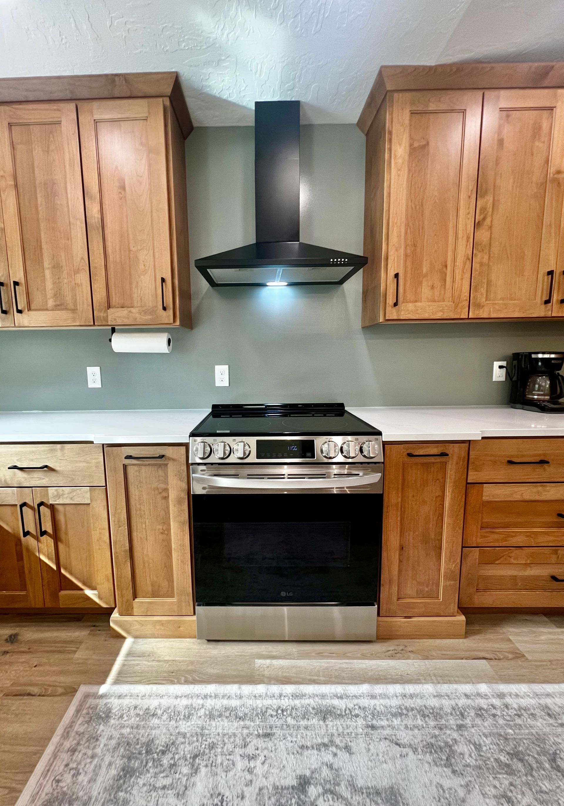 A kitchen with light wood cabinets, sage green walls, a stainless steel gas range, and a black chimney-style hood.