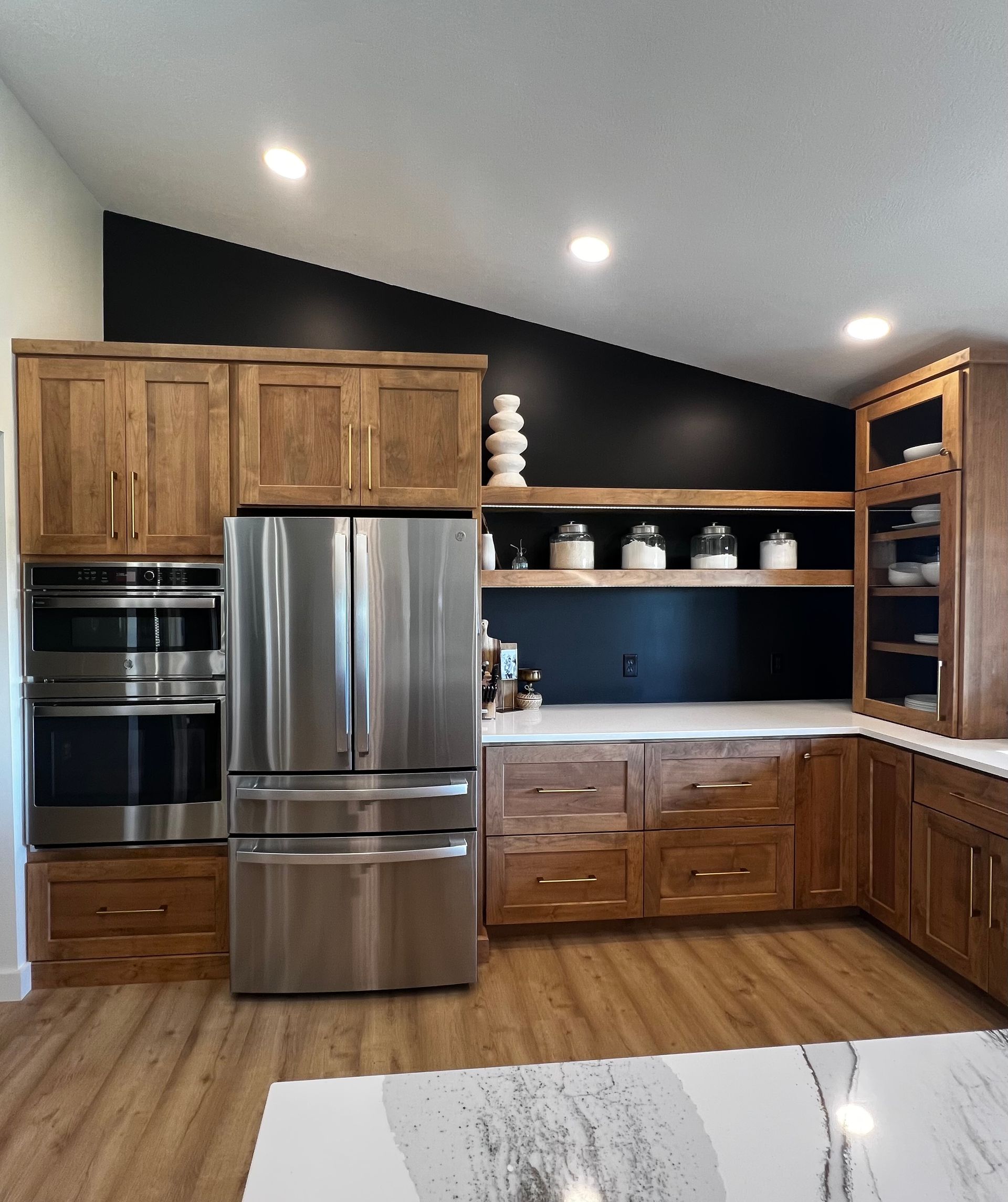 A modern kitchen features light wood cabinets, a stainless steel refrigerator, double ovens, and a black accent wall.