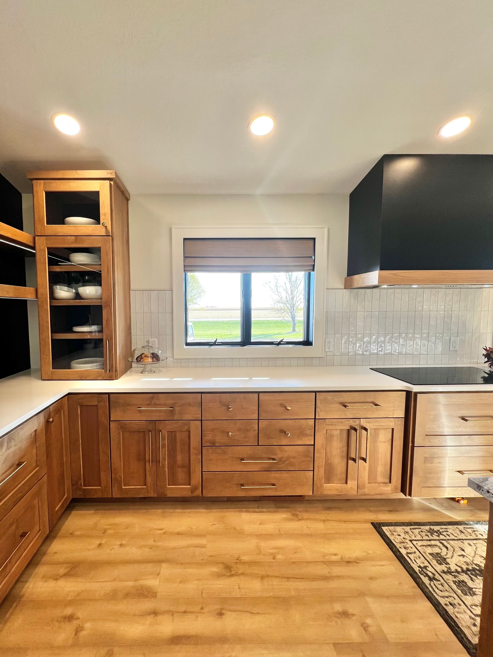 A modern kitchen features light wood cabinets, white countertops, white subway tile backsplash, and a black range hood.