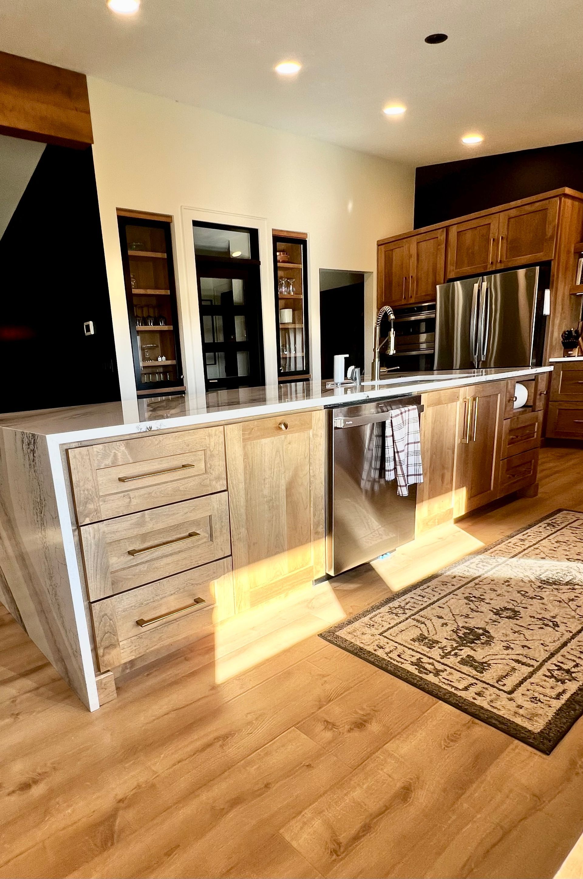 A modern kitchen island with light wood cabinets and white countertops, featuring a dishwasher and an area rug nearby.
