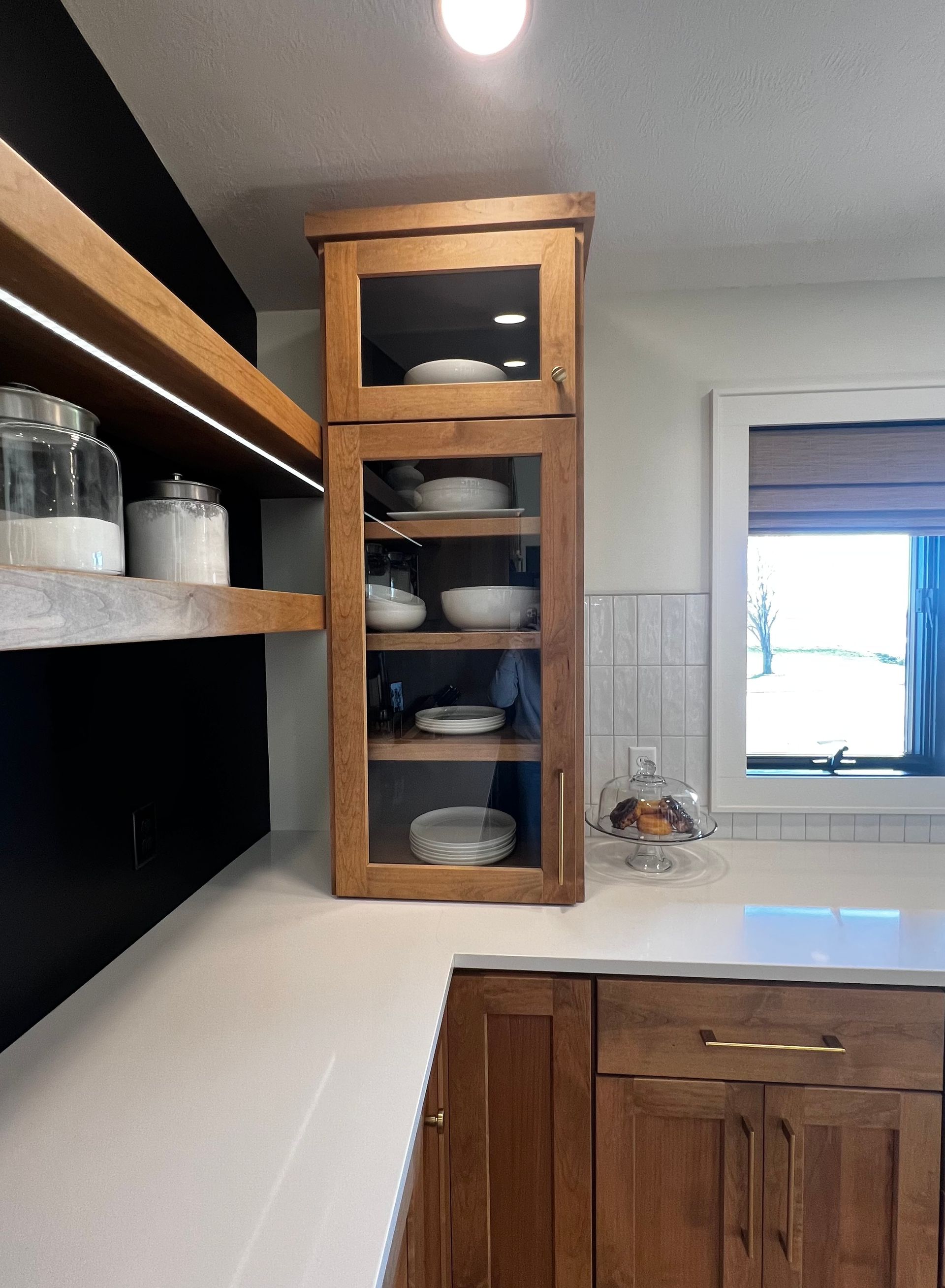 A kitchen corner with wooden cabinets, a glass-front display cupboard, open shelving on a black wall, and white counters.