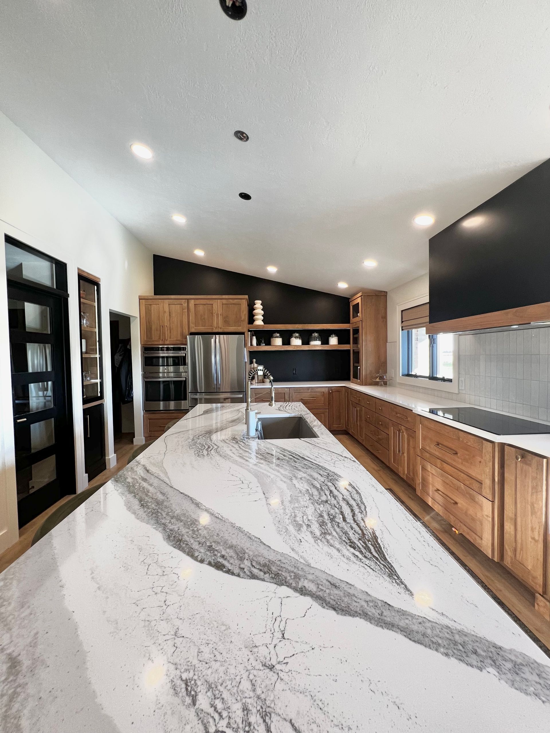 Modern kitchen featuring a long island with a white and grey marble countertop, wood cabinets, and black wall accents.