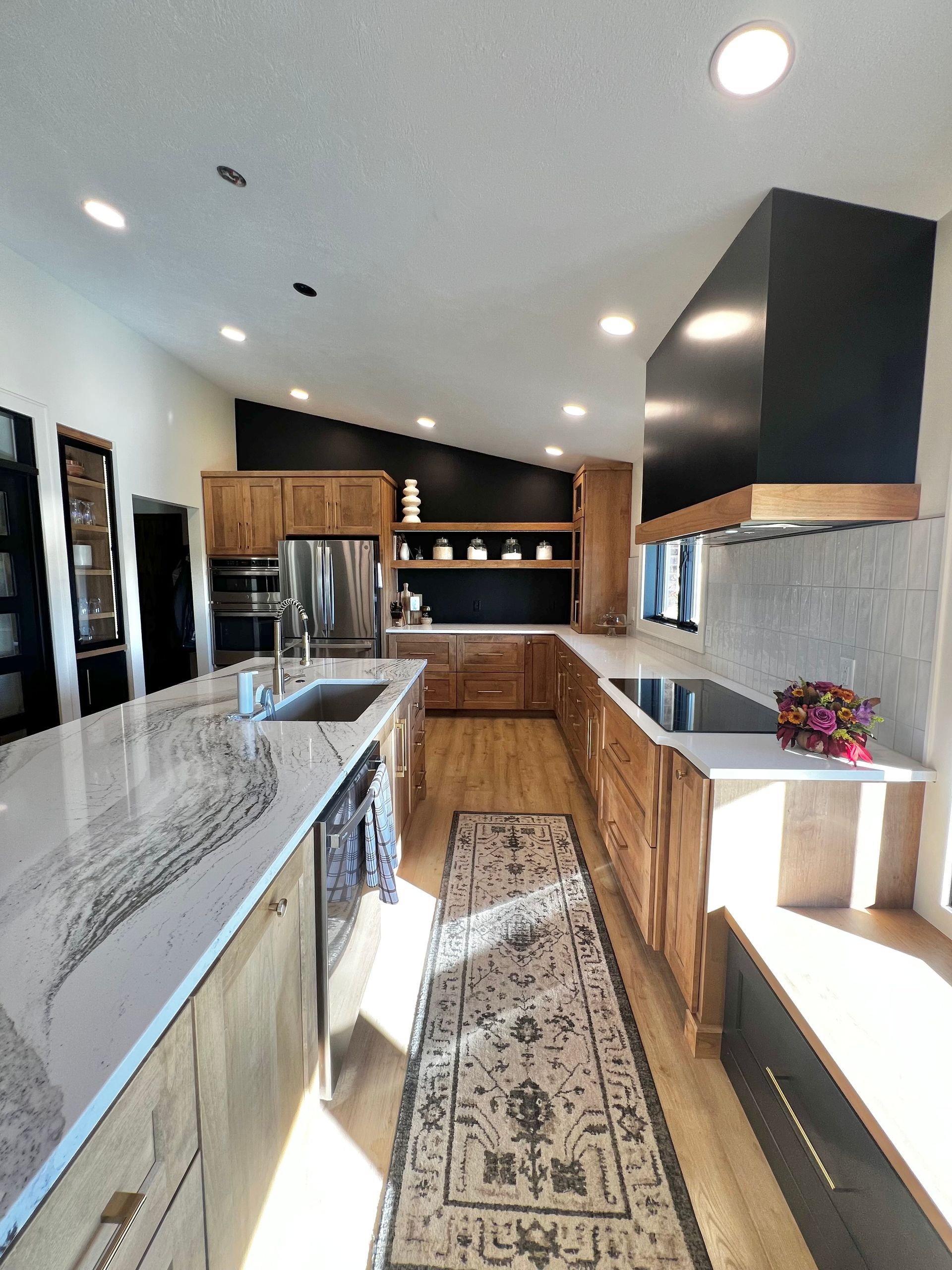 A modern kitchen features wood cabinets, white marble countertops, a black range hood, and a long patterned runner rug.