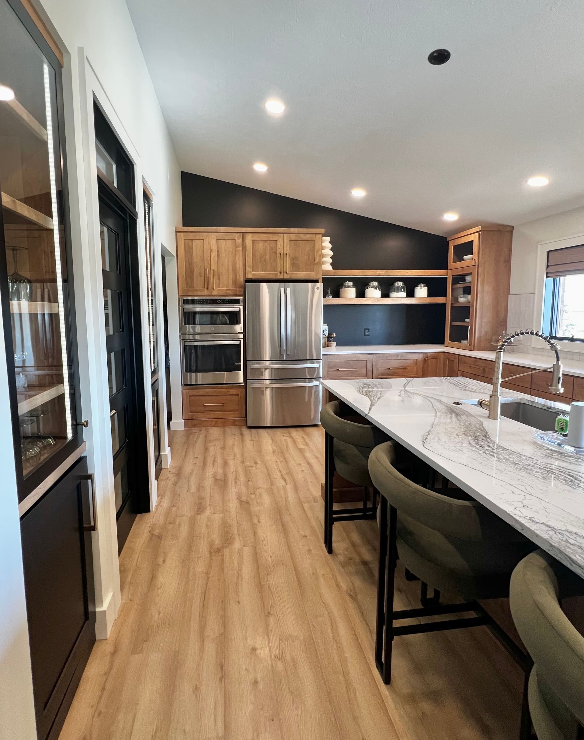Modern kitchen with natural wood cabinets, stainless steel appliances, a black accent wall, and a large marble island.