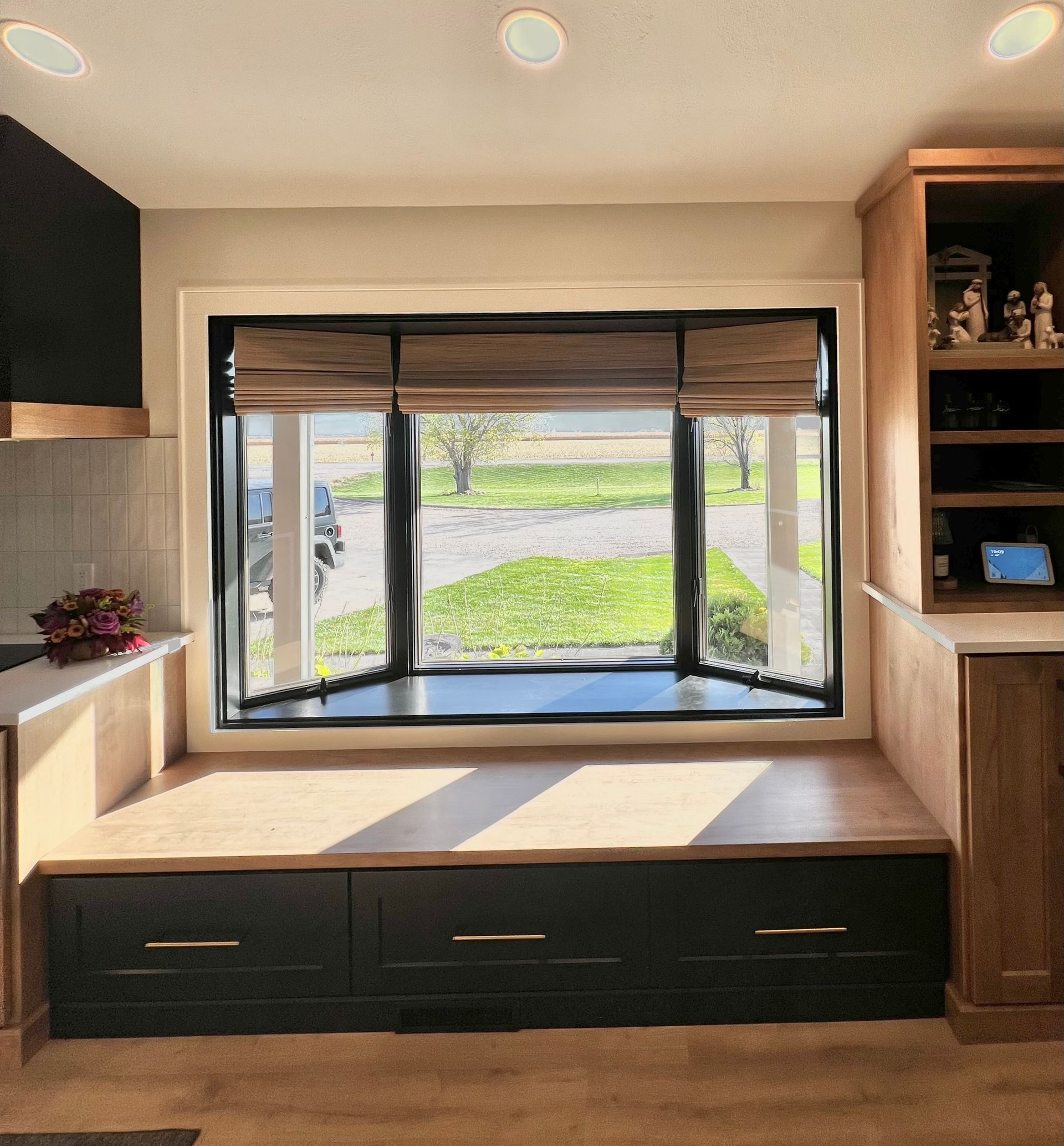 A bay window with wooden blinds, overlooking a yard, features a built-in bench with three black drawers beneath it.