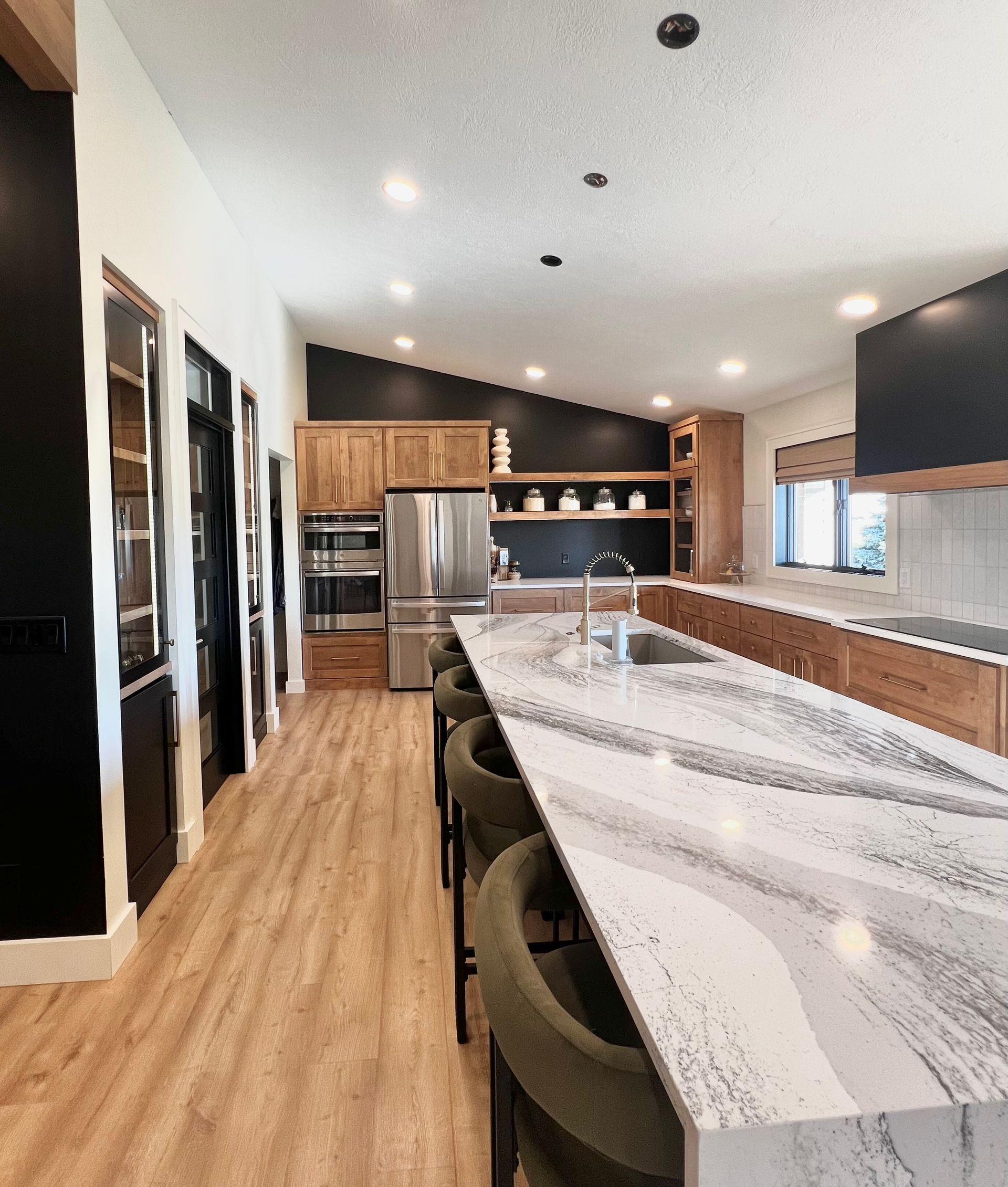 A spacious, modern kitchen with wood cabinets, black accents, a long marble-patterned island, and green bar stools.