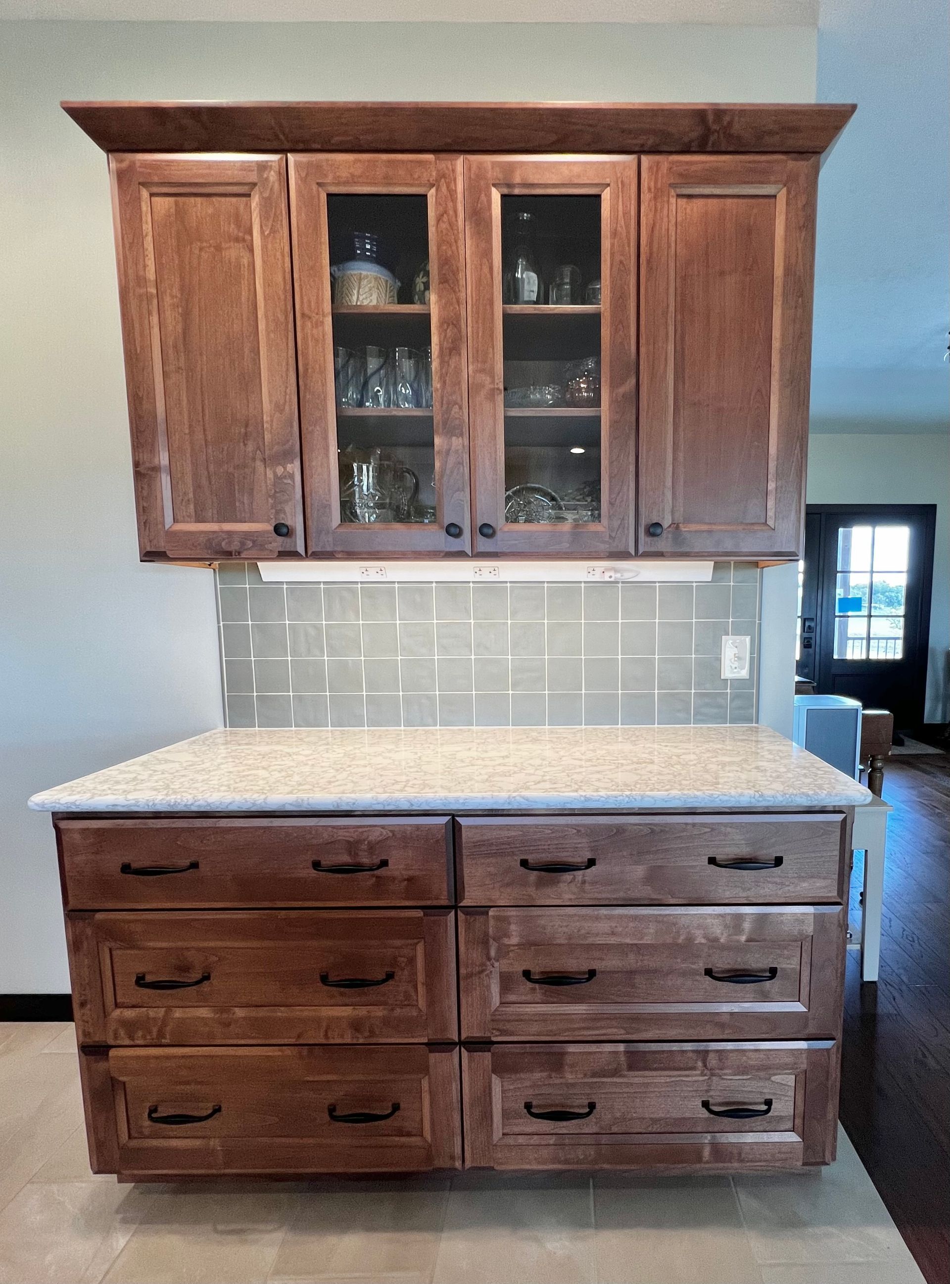Dark wood kitchen cabinets with glass doors sit above a speckled countertop and a six-drawer base unit with a tile wall.