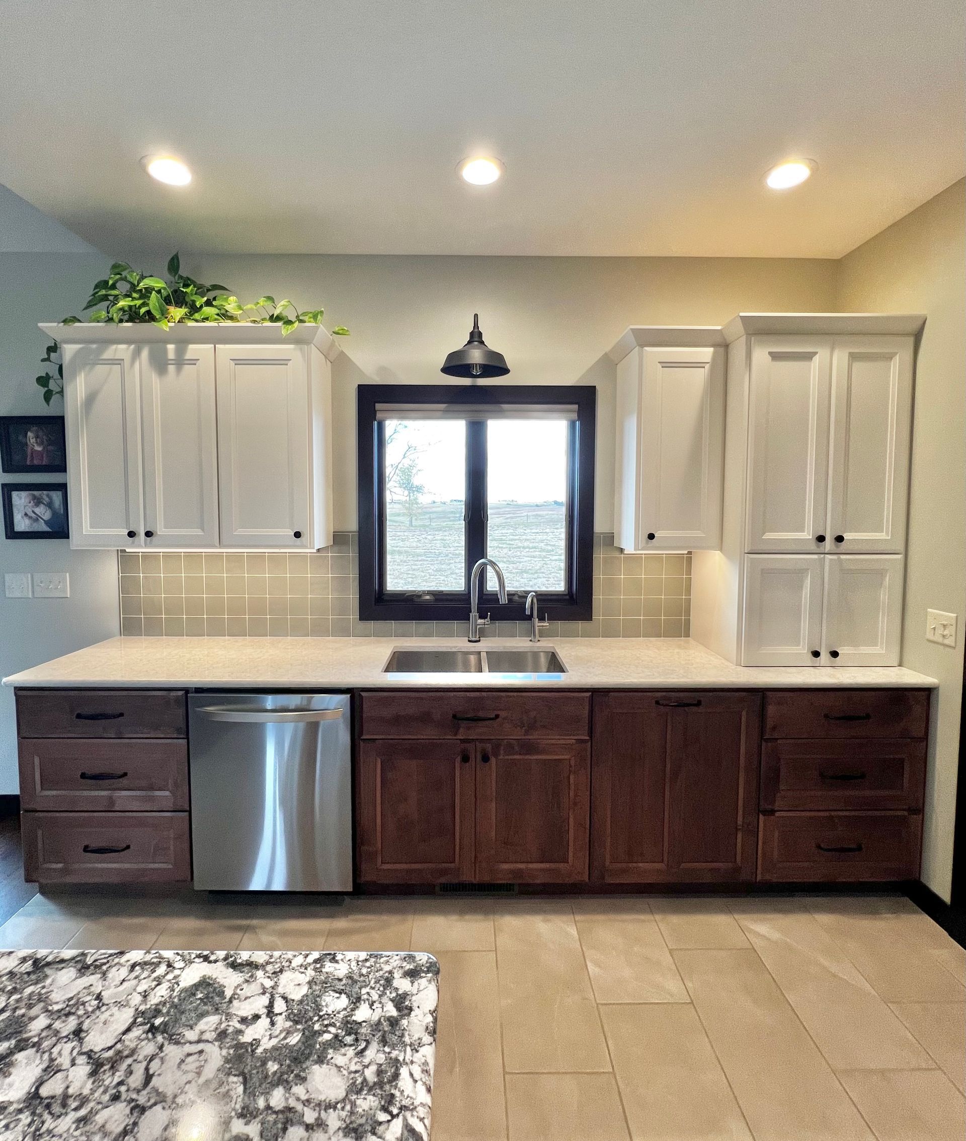 A kitchen with white upper cabinets, dark wood lower cabinets, a stainless dishwasher, and a window above the sink.