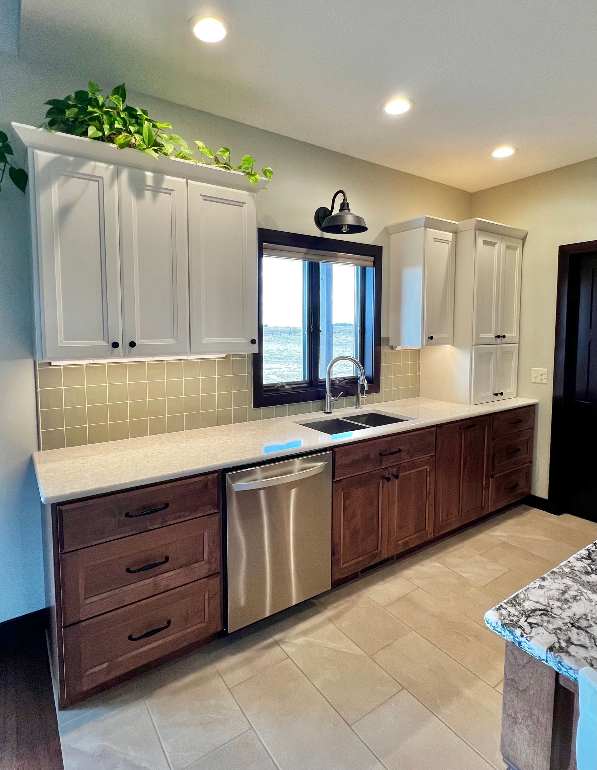 A kitchen with white upper cabinets, dark wood lower cabinets, light countertops, a stainless dishwasher, and a window.