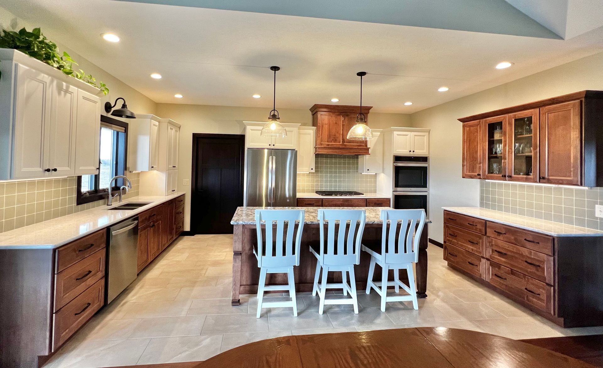 A kitchen with white upper cabinets, wood lower cabinets, a central island with three light-blue chairs, and tile floors.