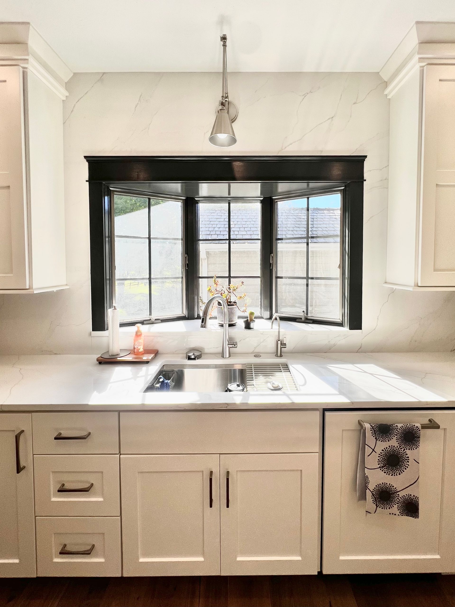 A kitchen sink area featuring a black-framed bay window, light cabinetry, white countertops, and a pendant light above.