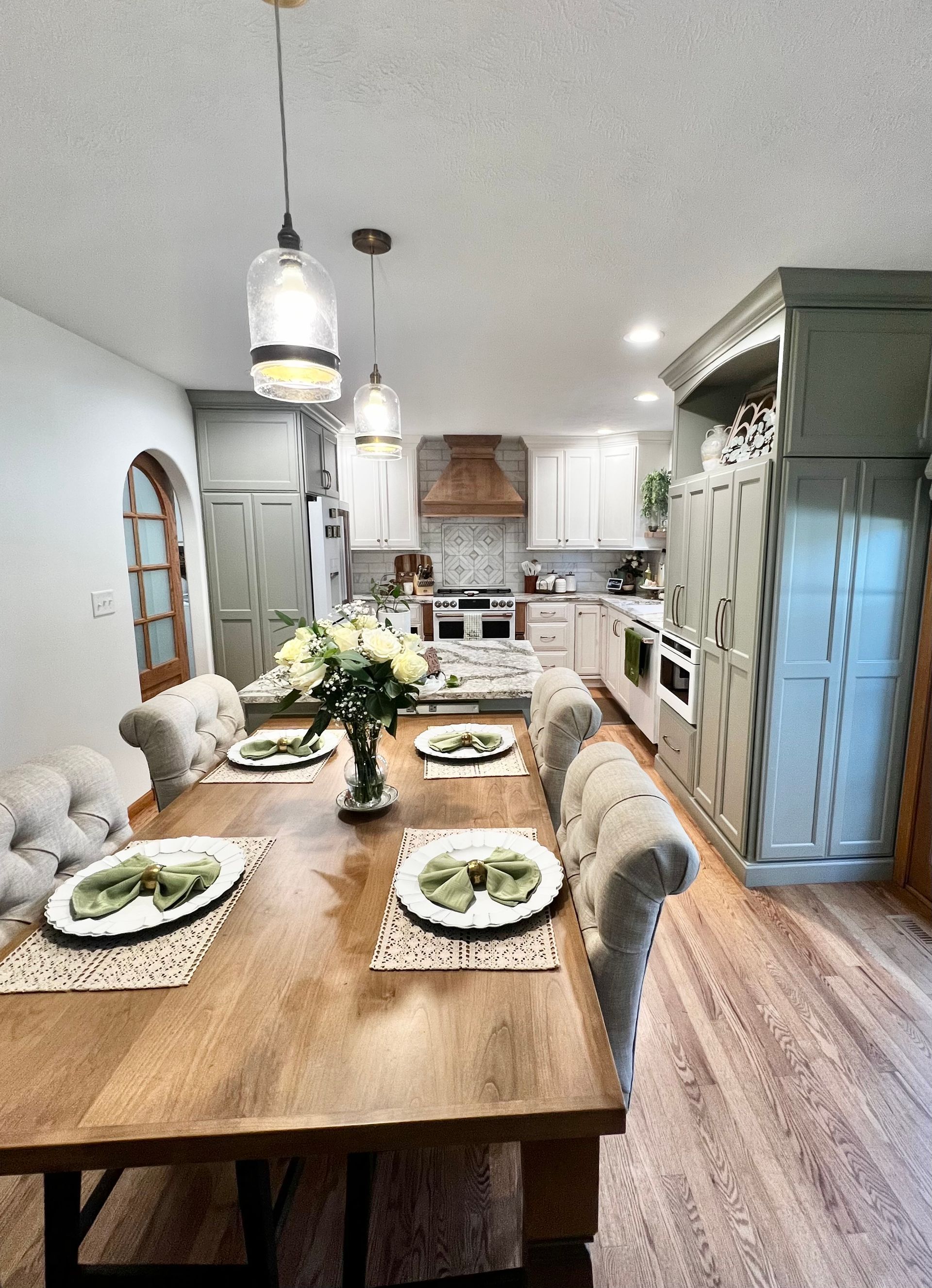 A wood dining table set for four in a brightly lit kitchen with cream-colored tufted chairs, light wood floors, and cabinets.