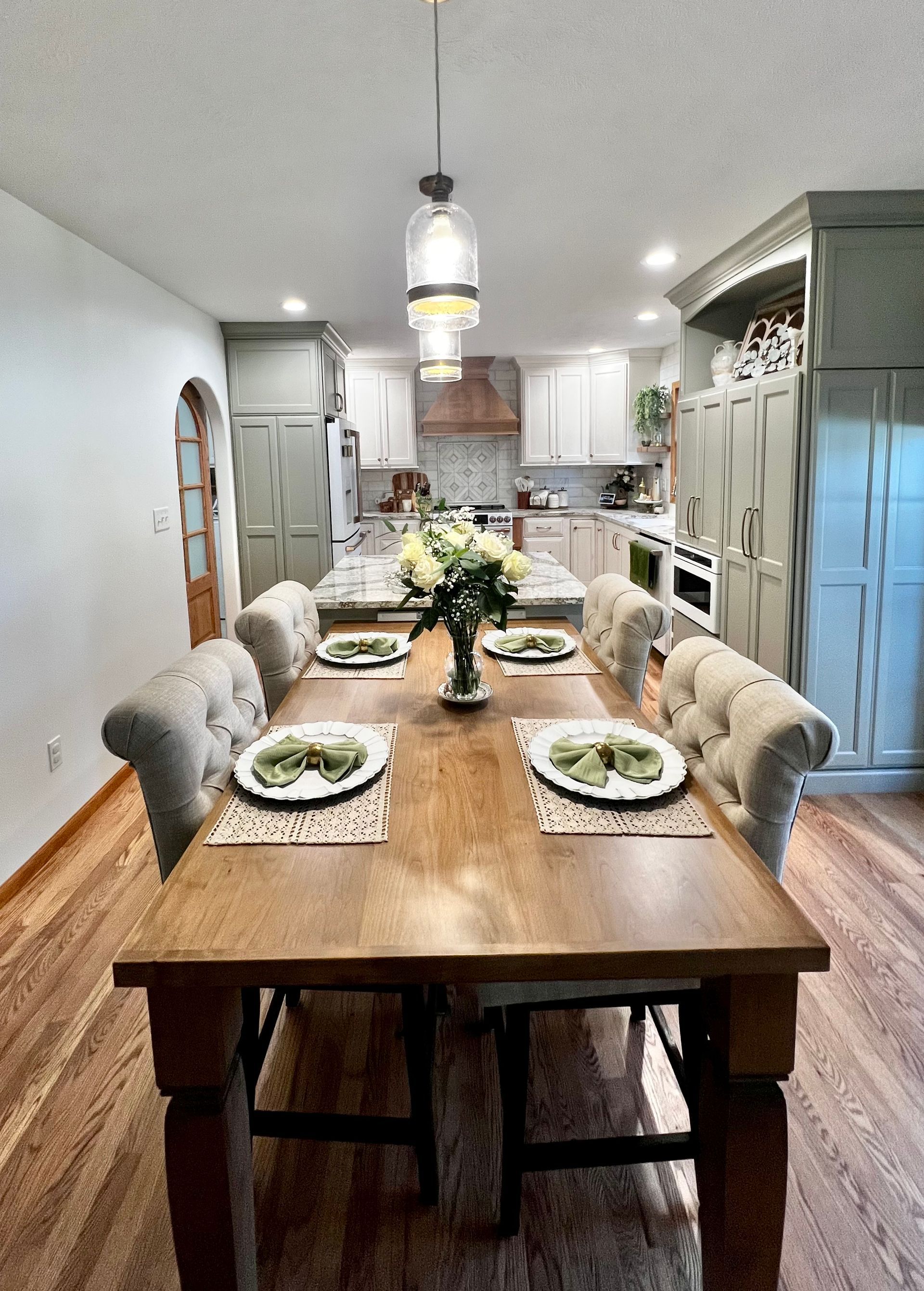 A wooden dining table with upholstered chairs sits in front of a white kitchen with green cabinetry and a pendant light.