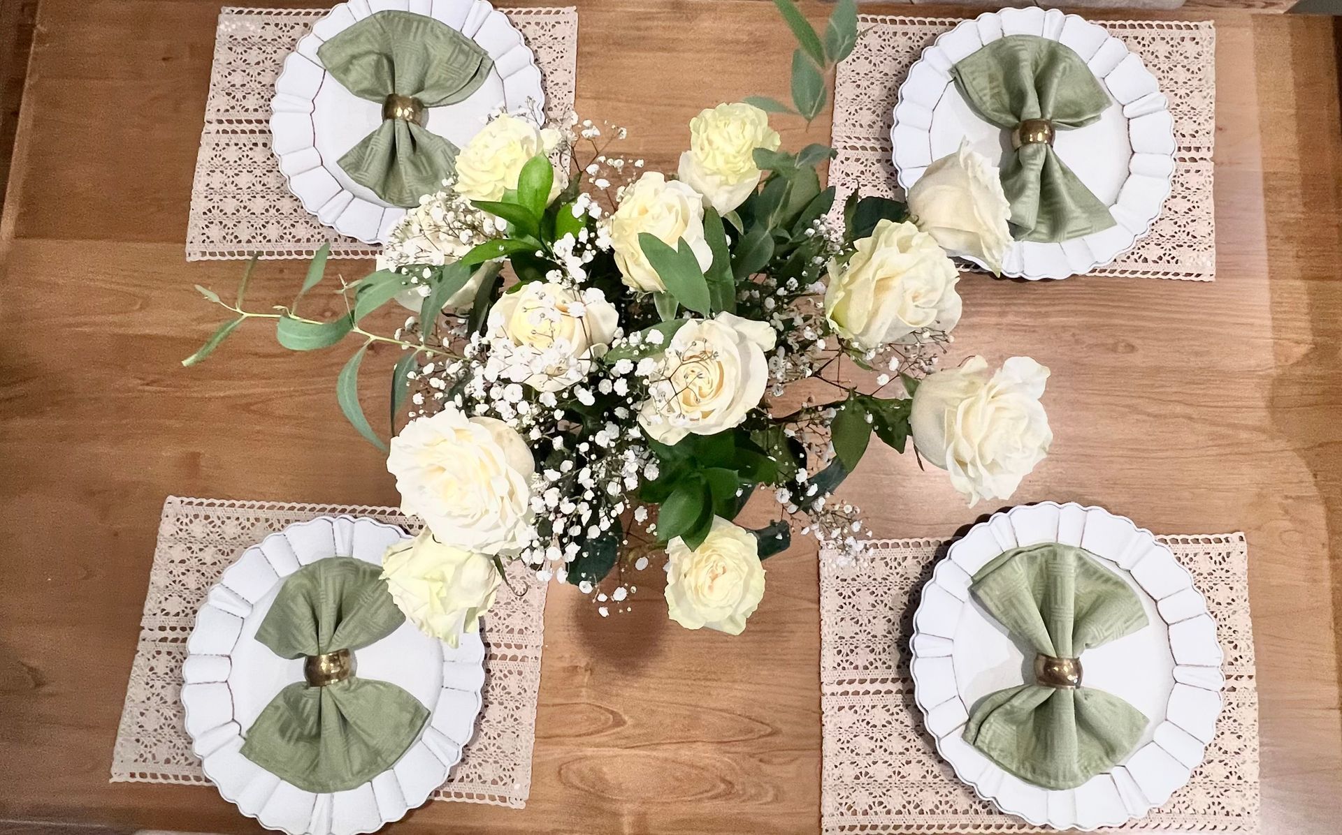 An overhead view of a wooden dining table with a central bouquet of white roses and four place settings with sage napkins.