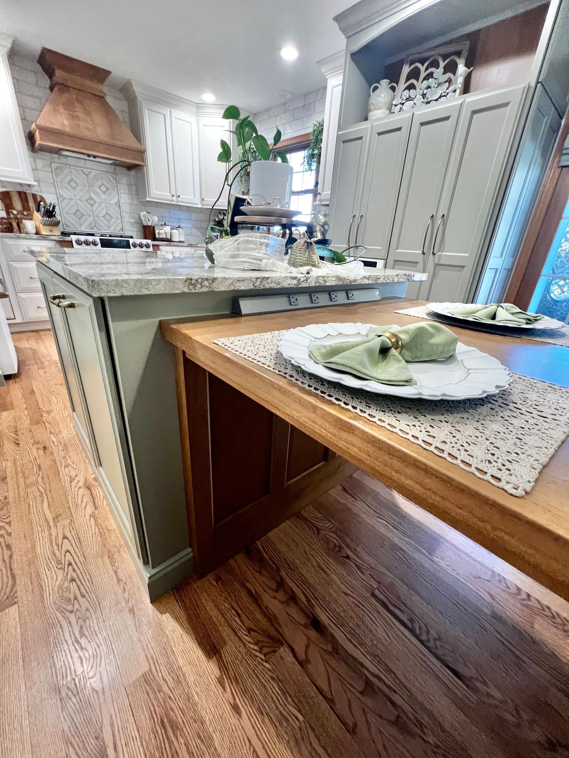 A kitchen island with a wooden table extension featuring place settings, set against green cabinetry and wood floors.
