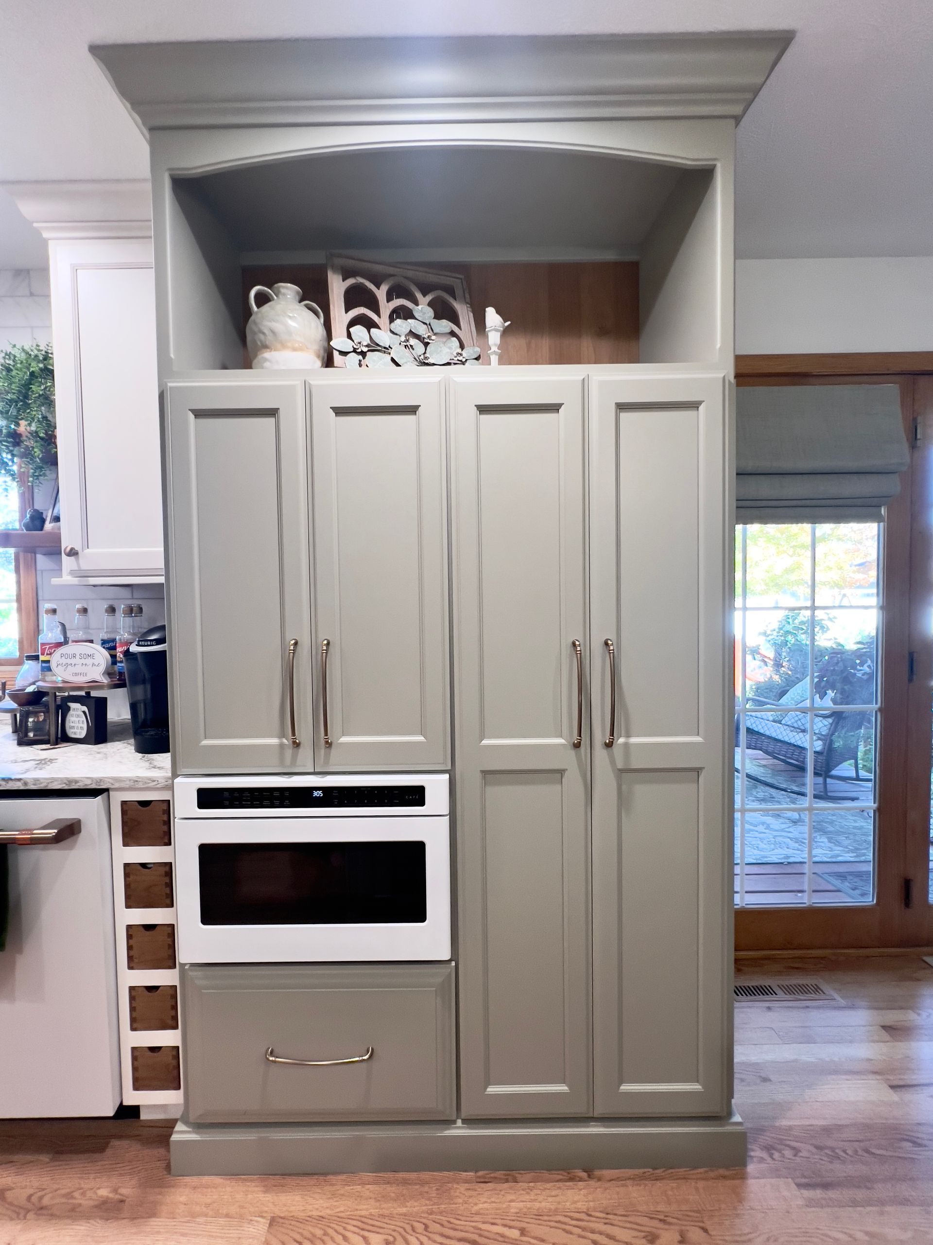Sage green floor-to-ceiling kitchen cabinet unit with a built-in microwave, drawer, and open display shelf at the top.