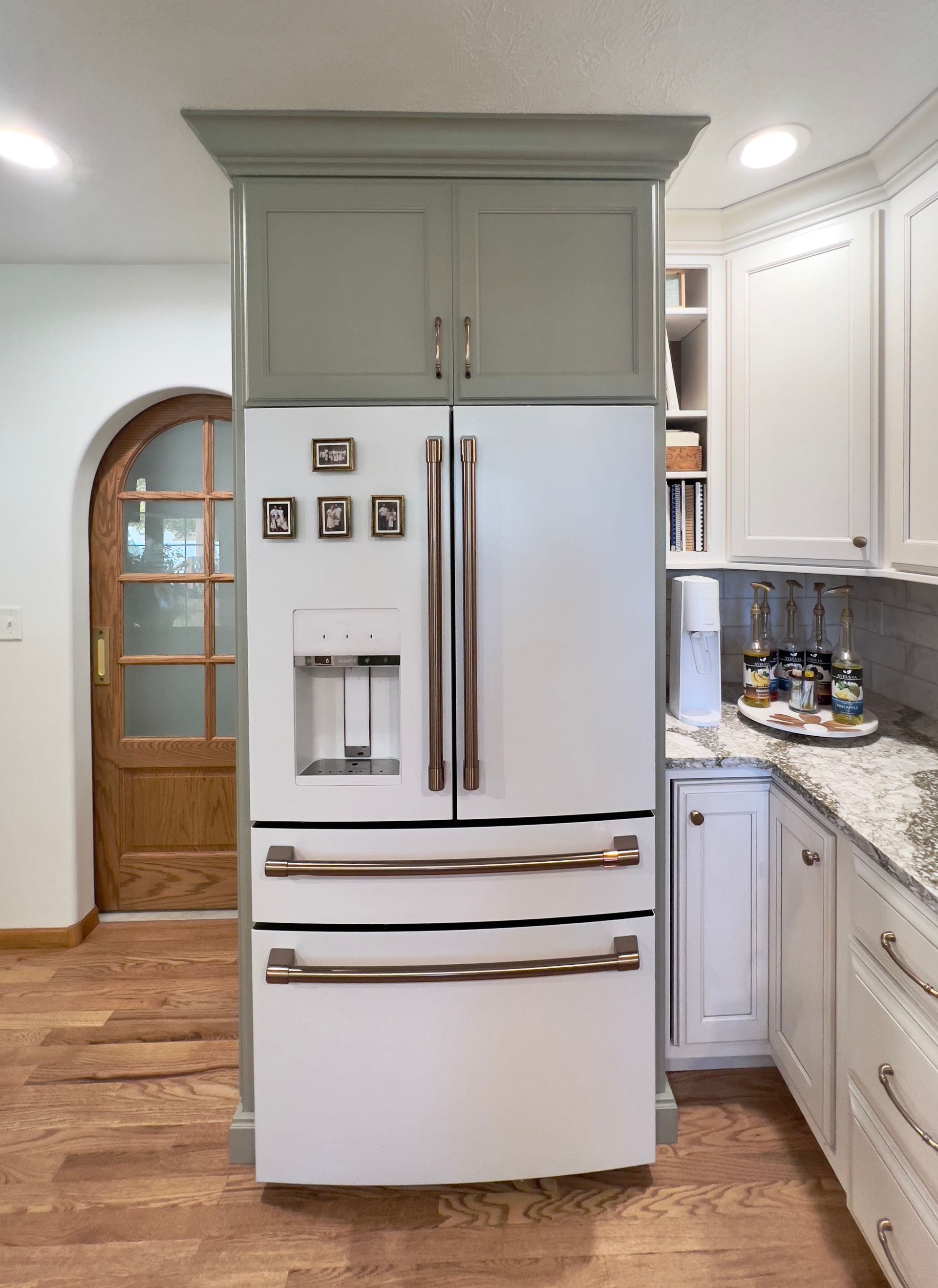 A white refrigerator with copper handles is set inside a gray cabinet frame in a kitchen with white cabinets and wood floors.