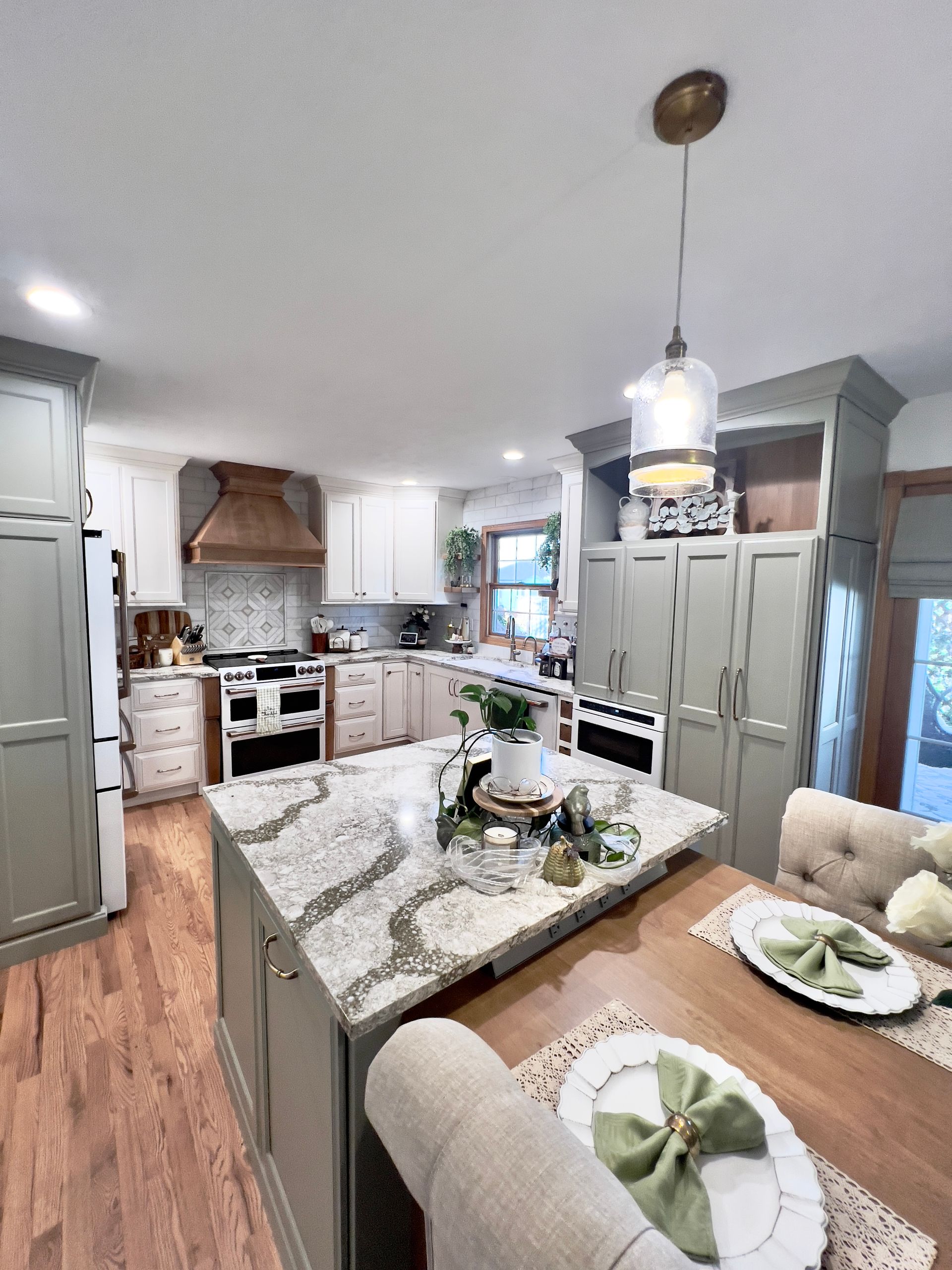 A kitchen with green cabinetry, white countertops, a wood range hood, a kitchen island, and a dining table with place mats.