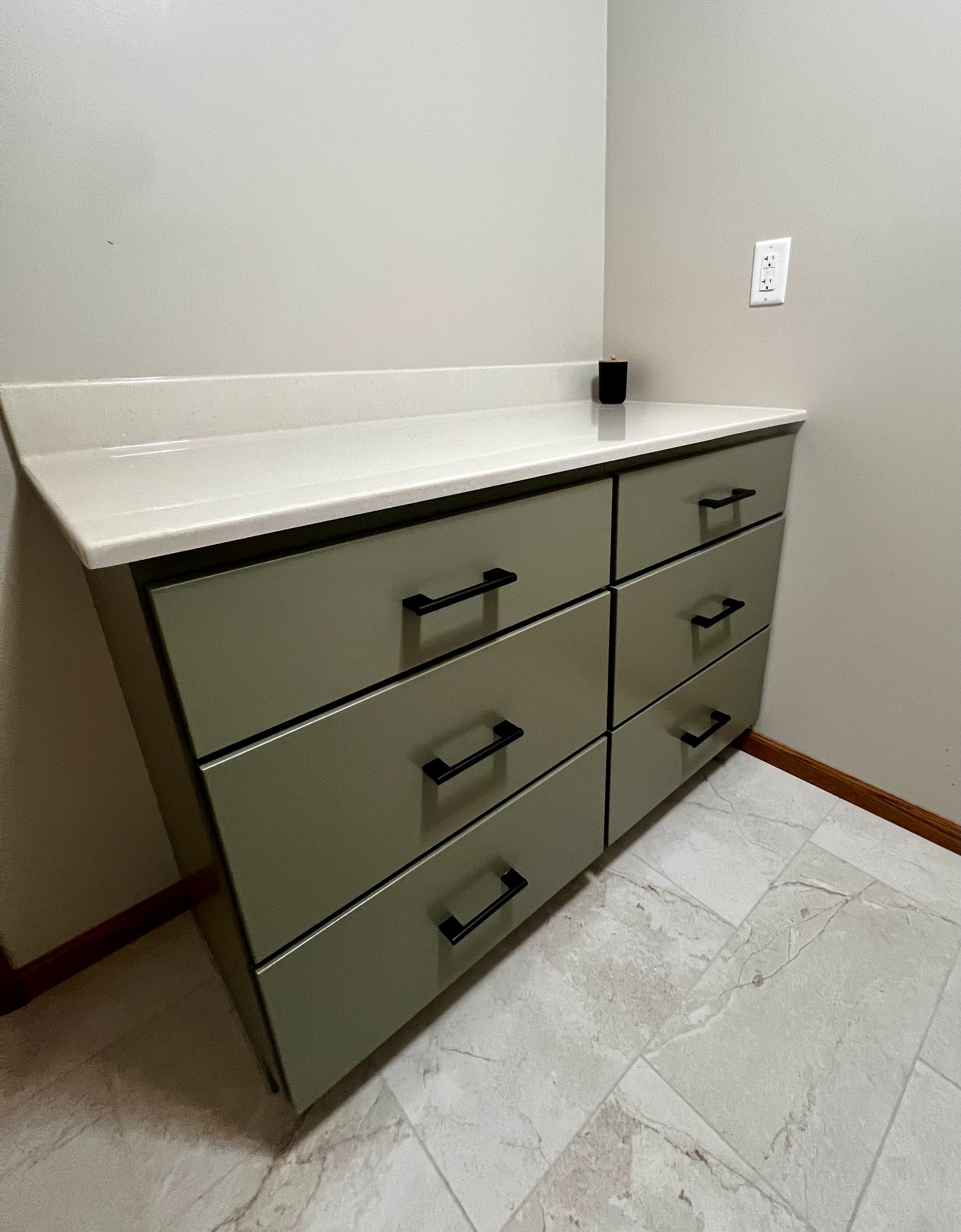 A sage-green six-drawer dresser with black hardware and a white countertop stands against a beige wall on a tiled floor.