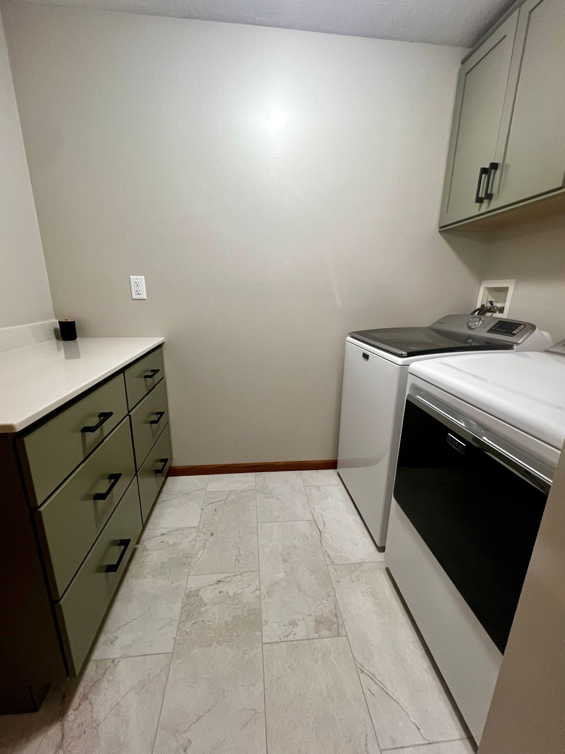 A laundry room with light-colored walls, a tiled floor, sage green cabinets, and a side-by-side washer and dryer.