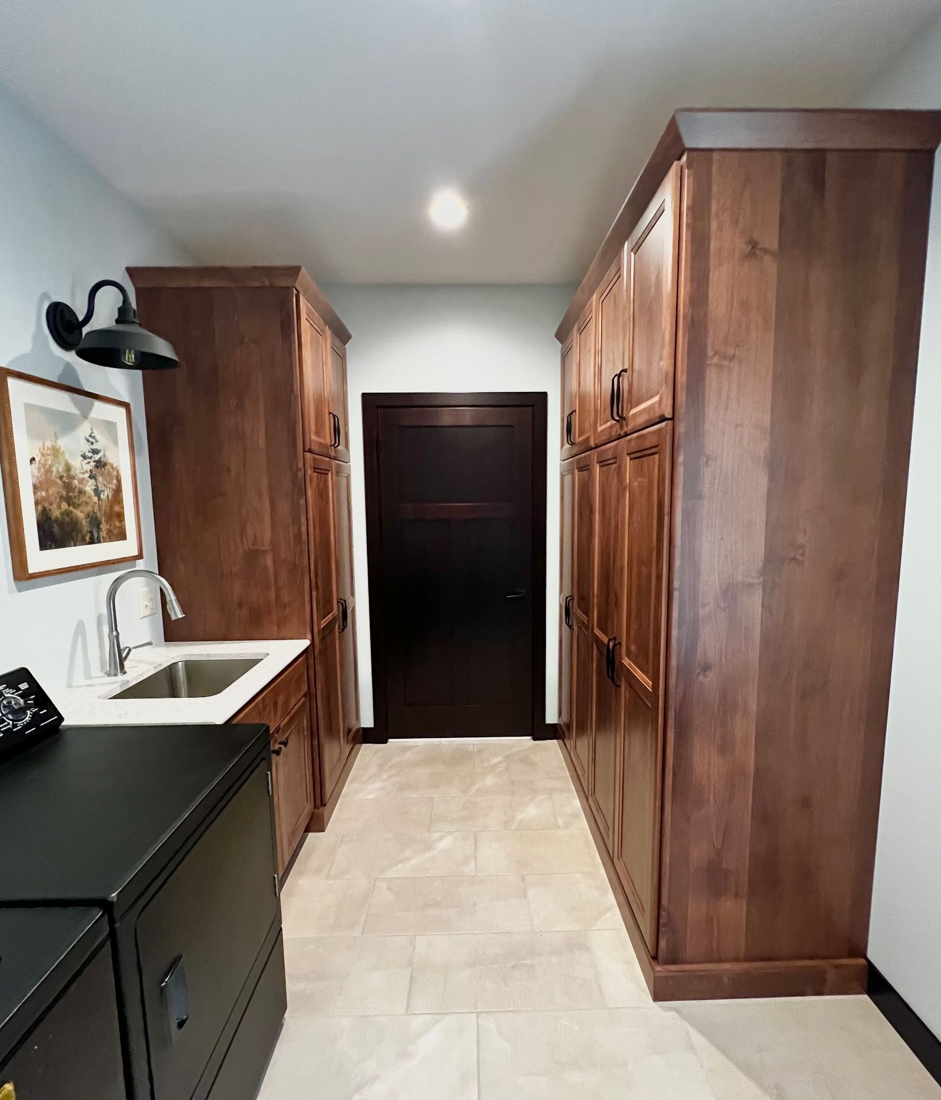 A laundry room features dark wood cabinetry, a sink, tile floors, and a black door at the end of the hallway.