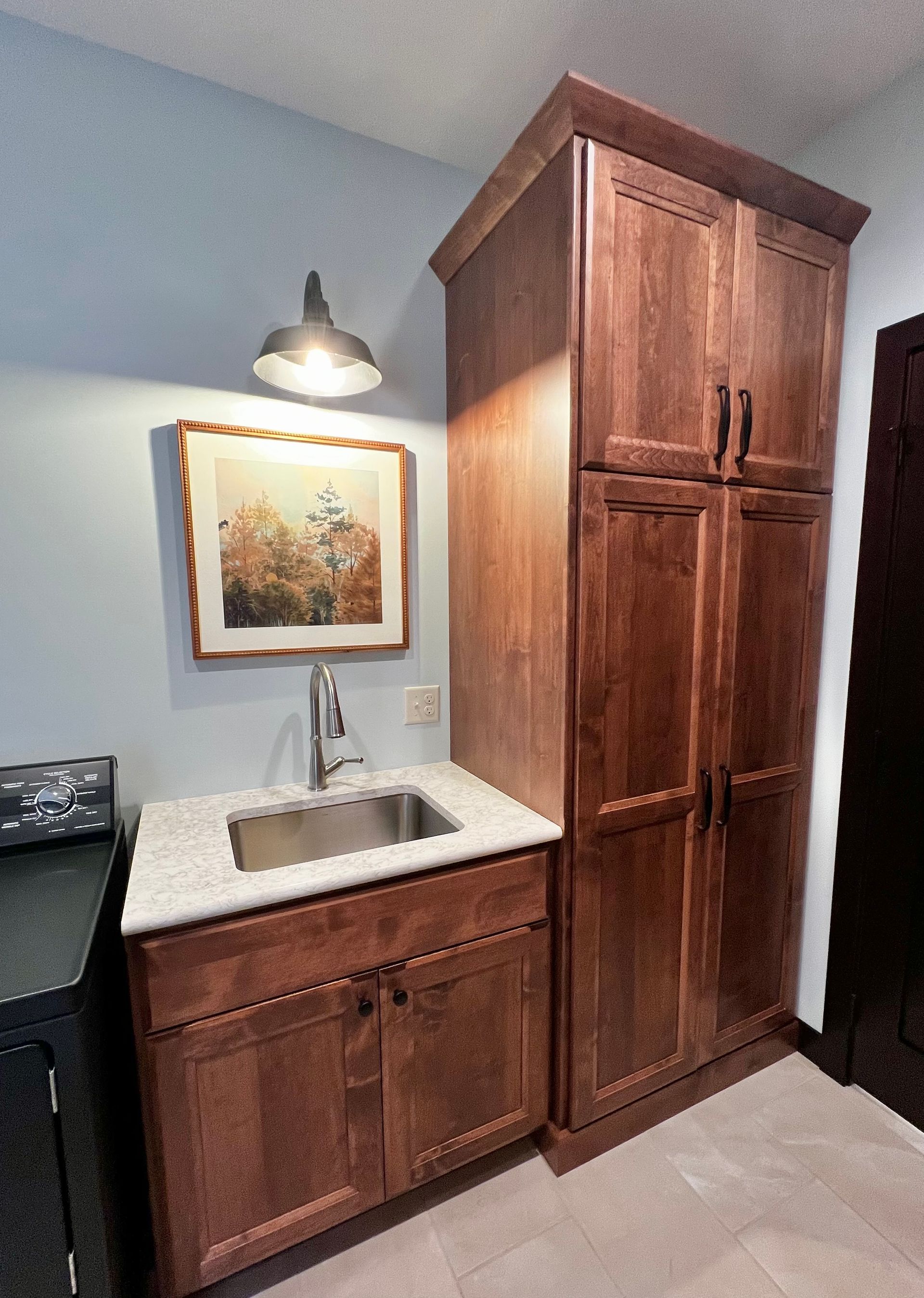 A sink and cabinet set with wooden cupboards, a white speckled countertop, a wall-mounted light, and framed artwork.