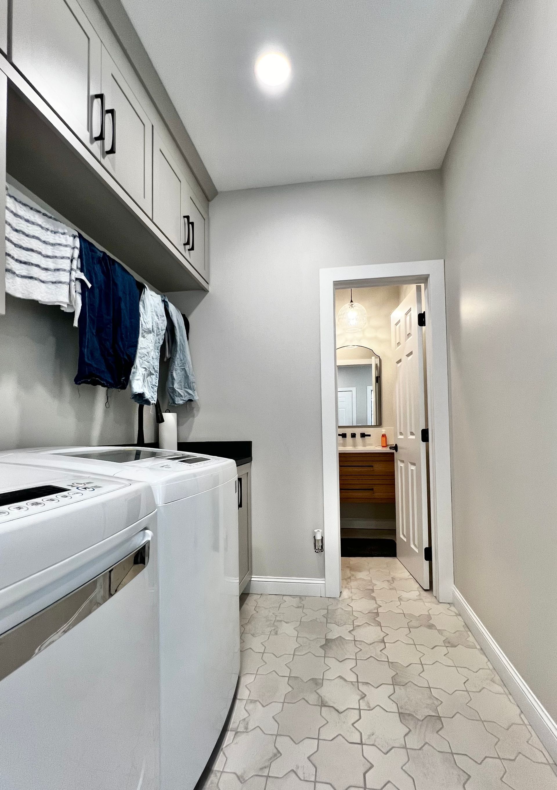 A laundry room with white appliances, gray cabinetry, hanging clothes, and a patterned tiled floor, leading to a bathroom.