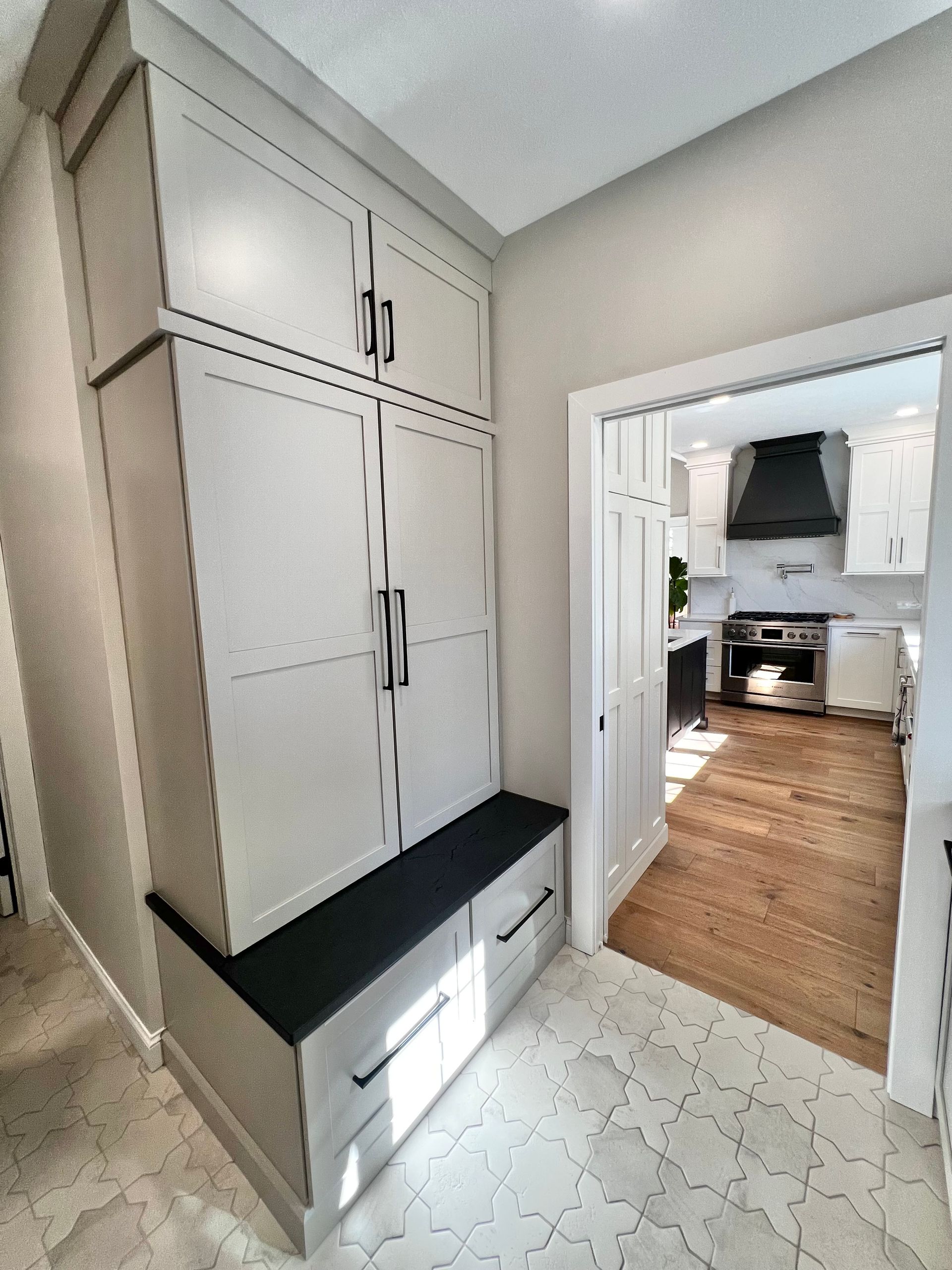 A tall gray storage cabinet with a built-in bench seat stands in a hallway, looking into a kitchen with hardwood floors.