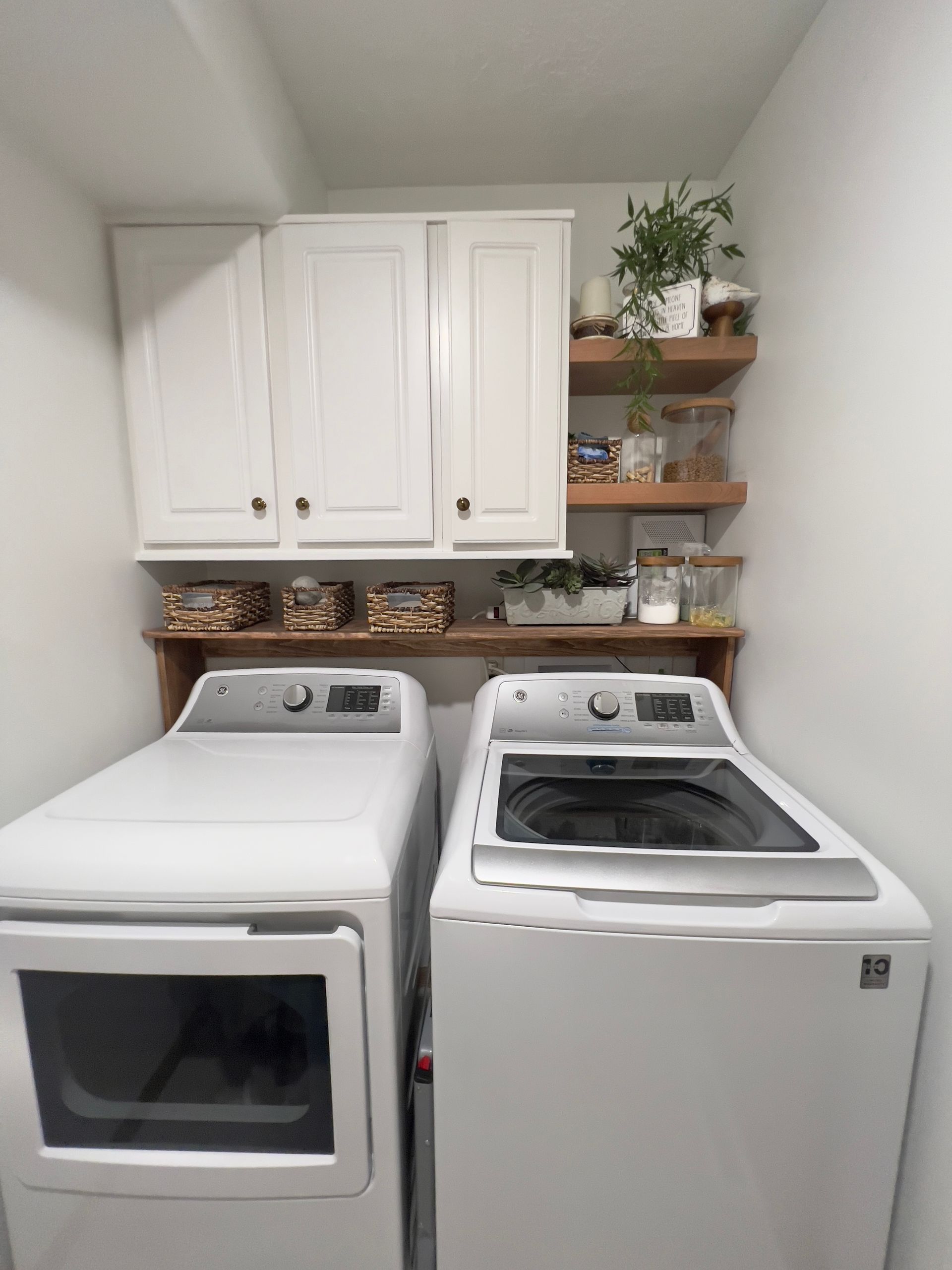 White washer and dryer under white cabinets and open wooden shelves with plants and baskets in a laundry room.