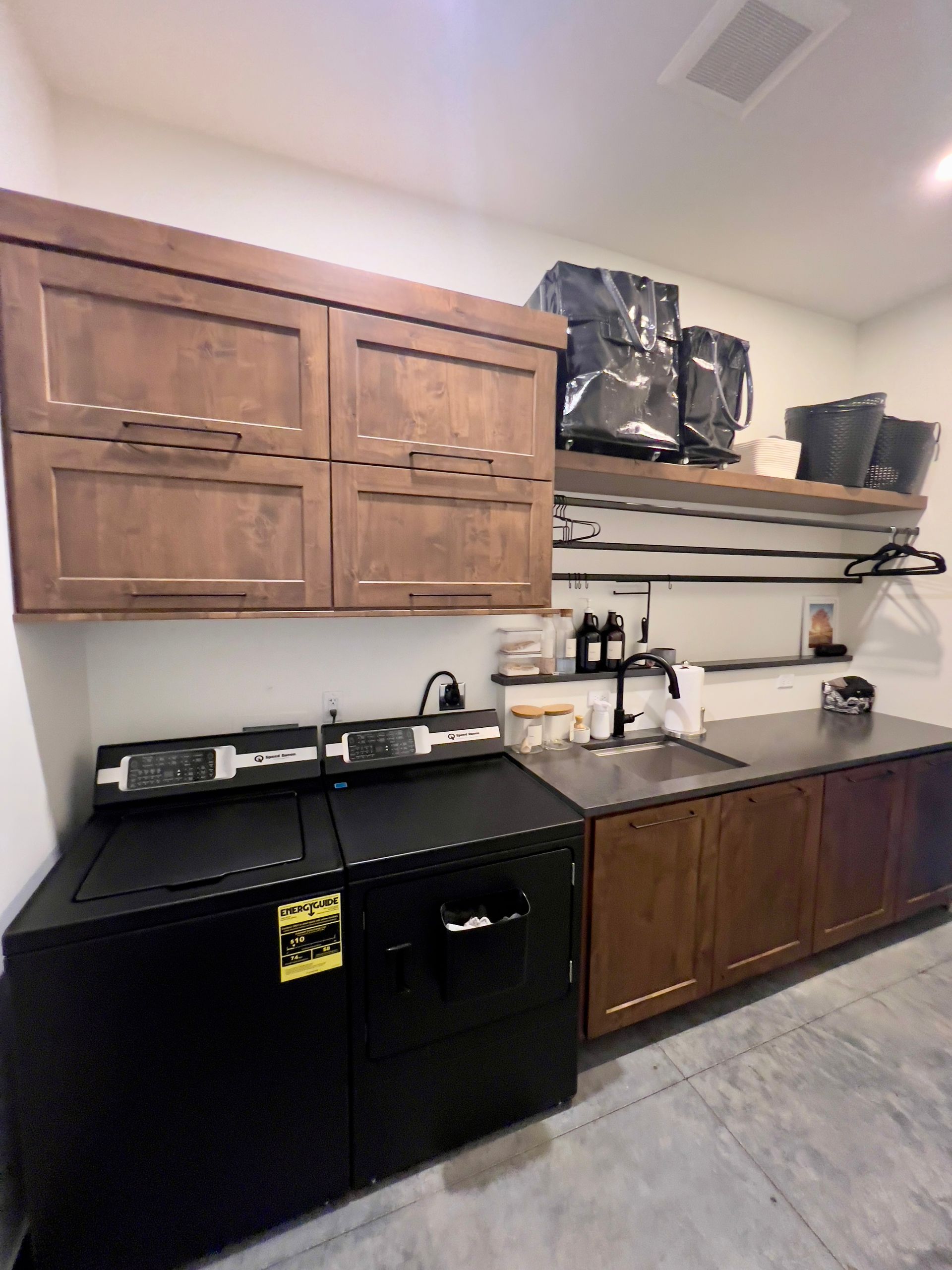 A laundry room with dark wood cabinets, a black washing machine, a dark countertop, a sink, and open shelving with bins.