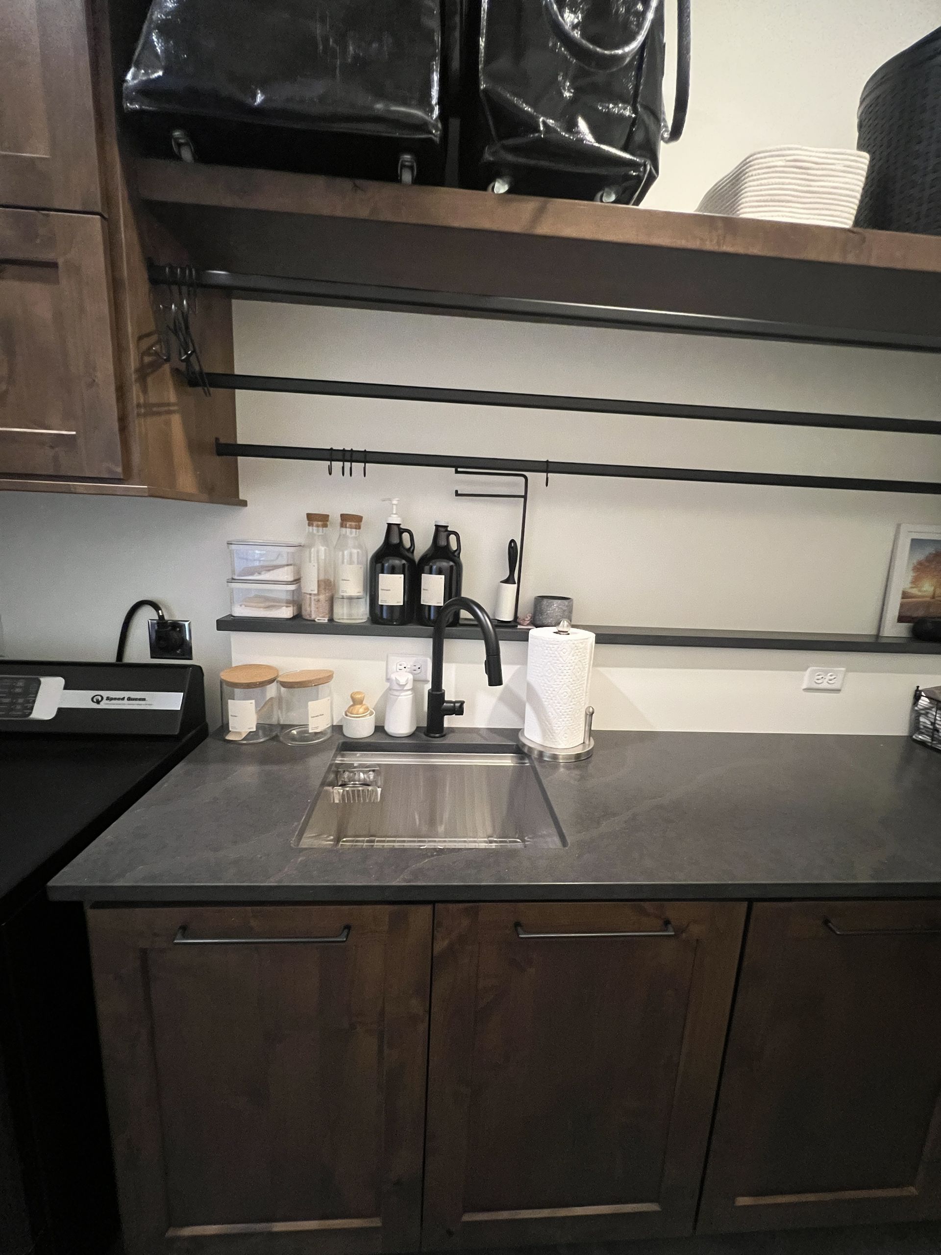 Laundry room sink with dark cabinets, grey countertops, and open shelving featuring black storage rods and containers.