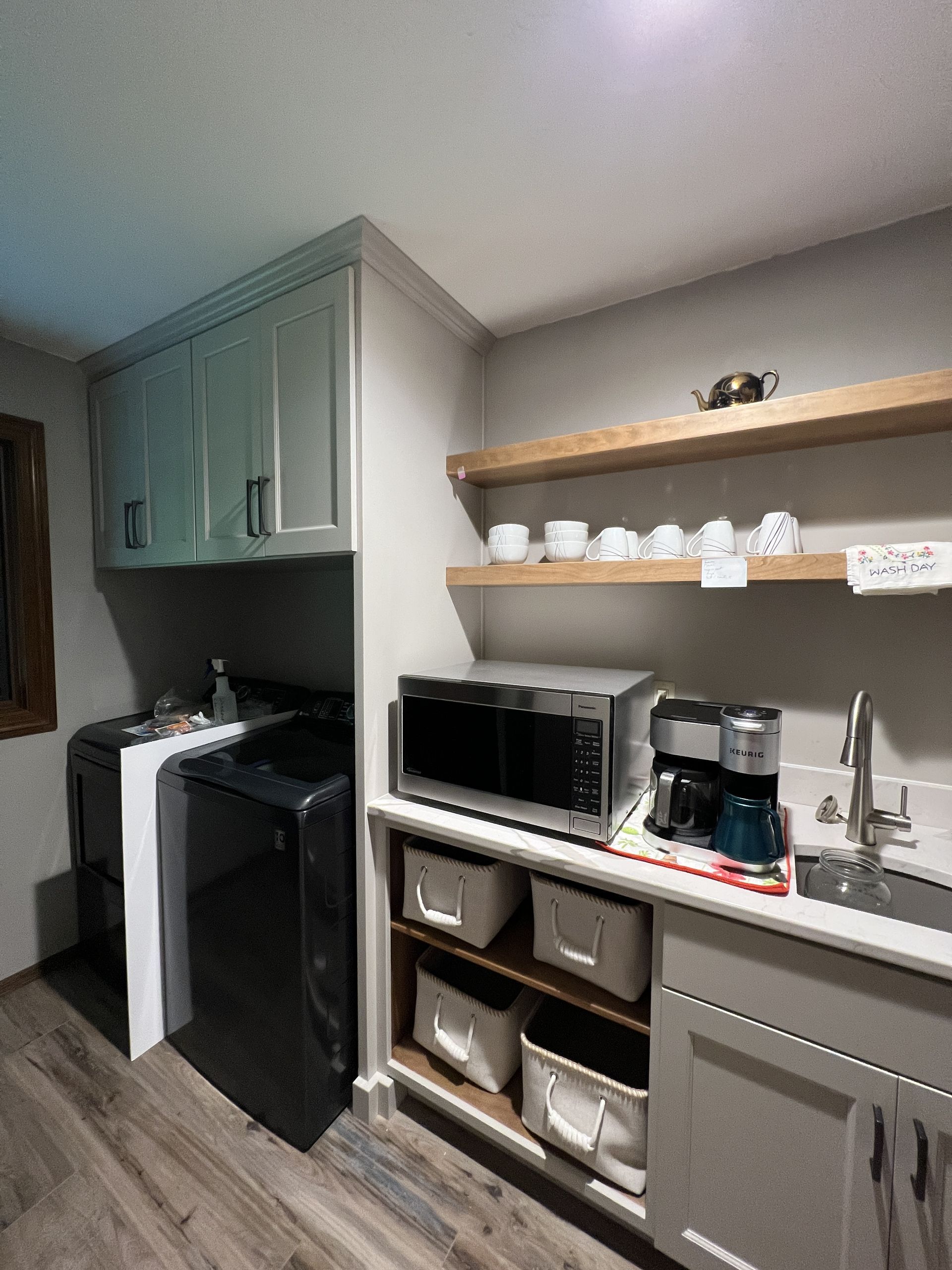 A kitchen nook with laundry appliances, cabinets, open shelving, a microwave, coffee maker, and sink on light countertops.
