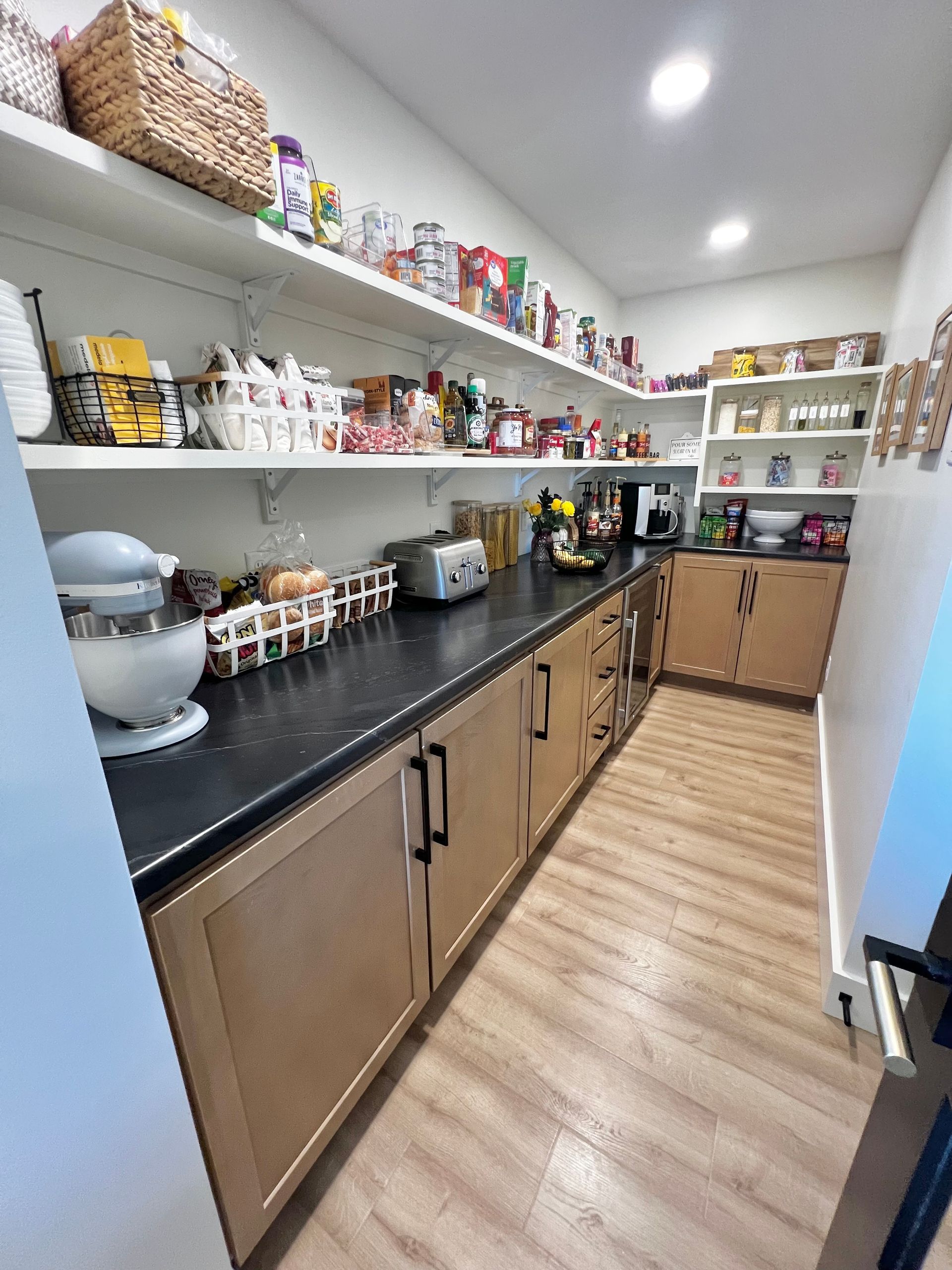 A walk-in pantry with light wood cabinets, dark countertops, white open shelving, and a stand mixer on the counter.