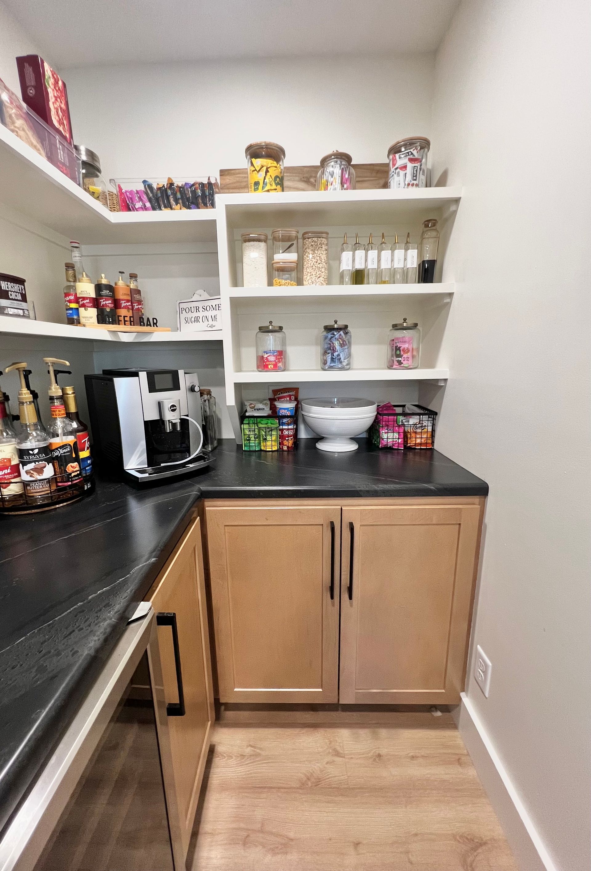 A corner pantry featuring light wood cabinets, black countertops, and open shelving filled with jars, snacks, and a machine.