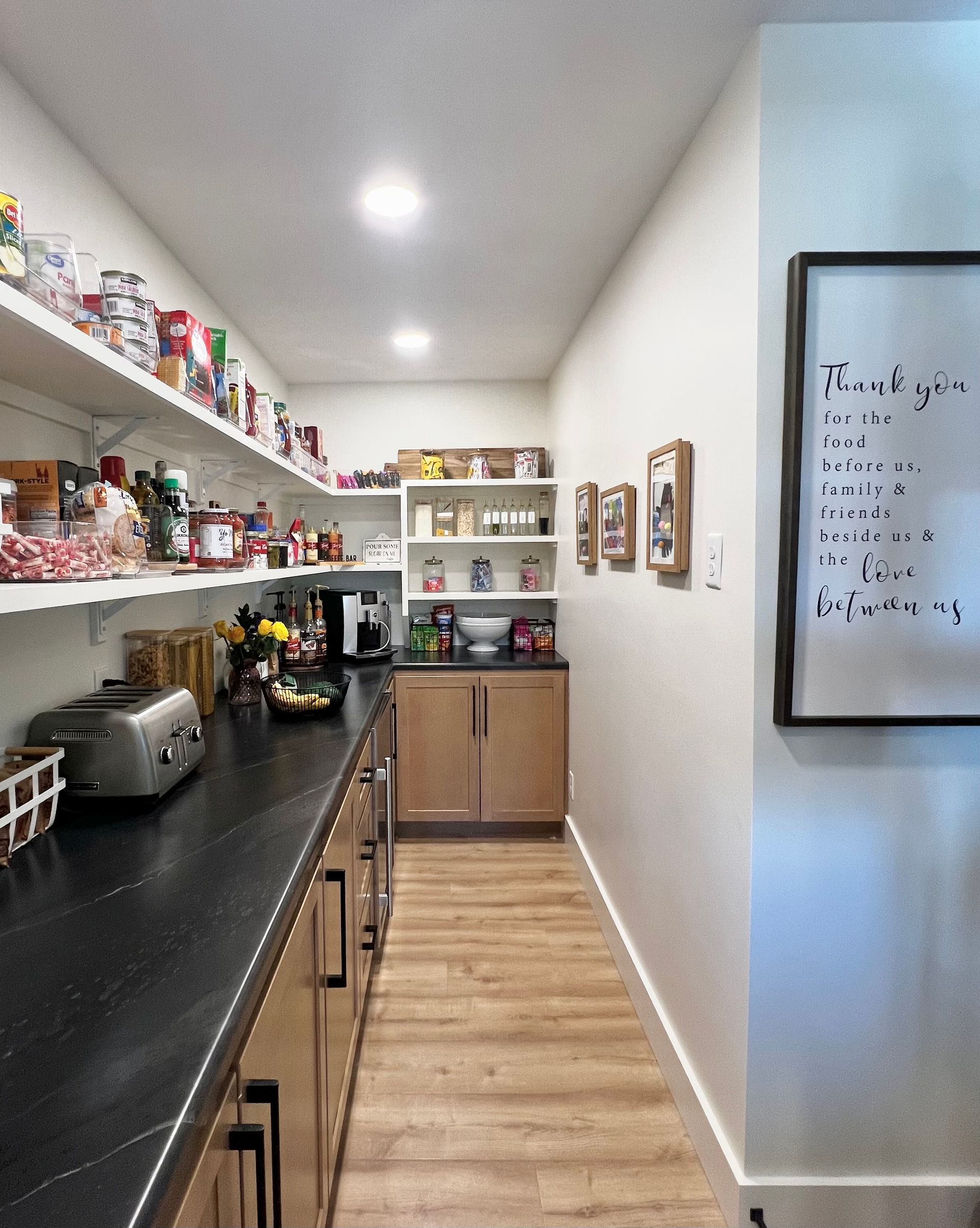 A pantry with wood cabinets, black countertops, open shelving filled with food, and a whiteboard on the wall.