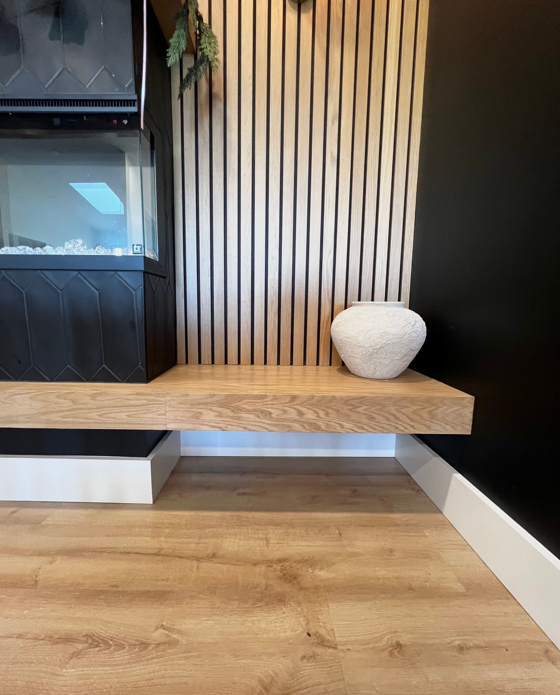 A light wood floating hearth shelf sits below a fireplace with vertical wooden slats against a dark wall.