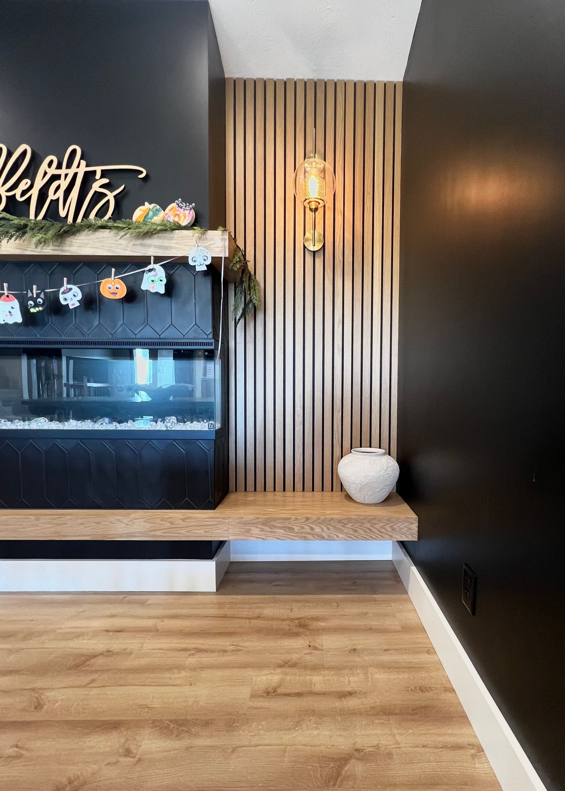 A modern fireplace with a wood-slat feature wall, a light wood mantel, and black walls in a room with wood flooring.
