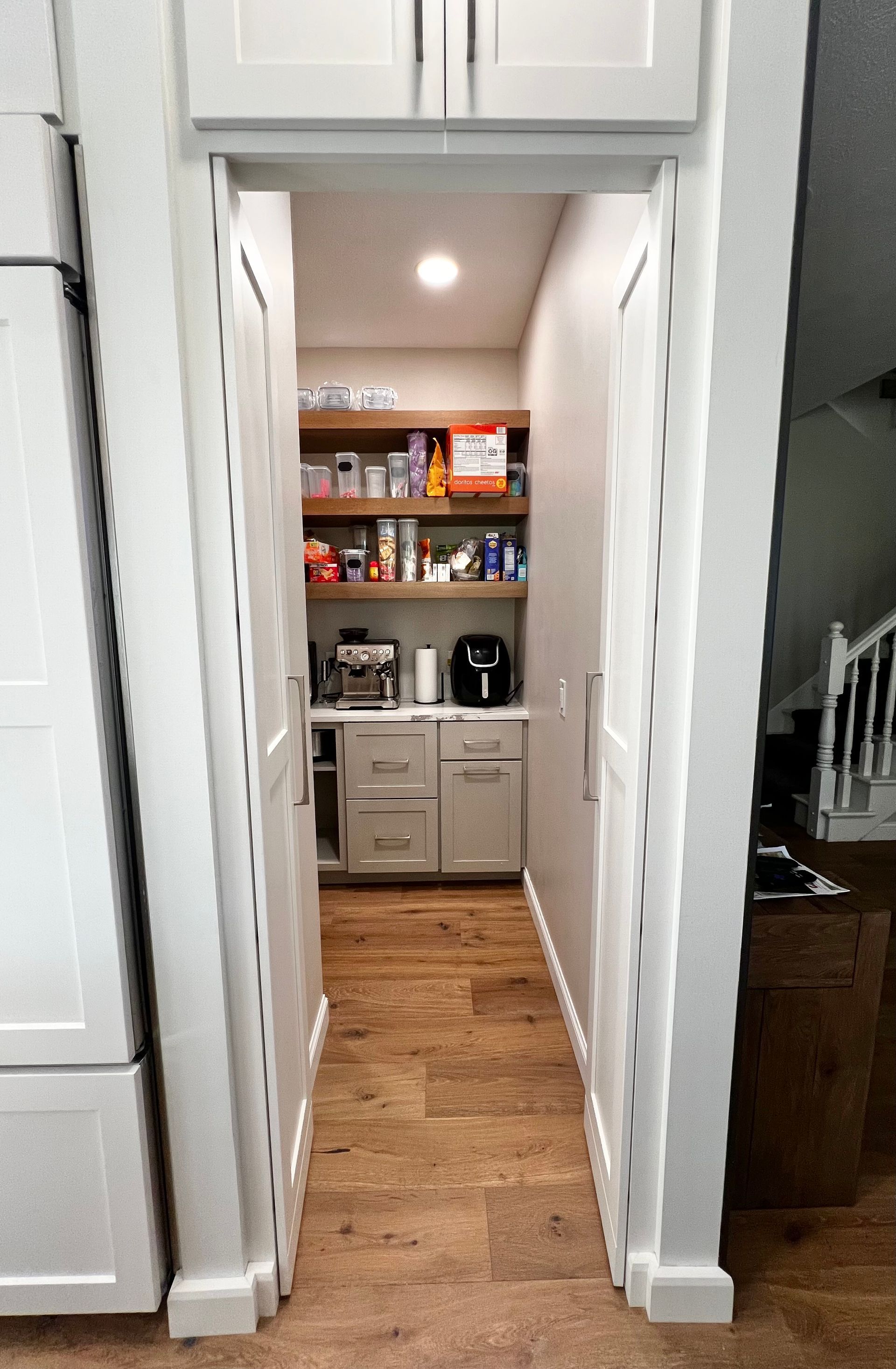 A narrow pantry with white cabinet doors, wooden shelves stocked with items, a countertop, and light wood flooring.