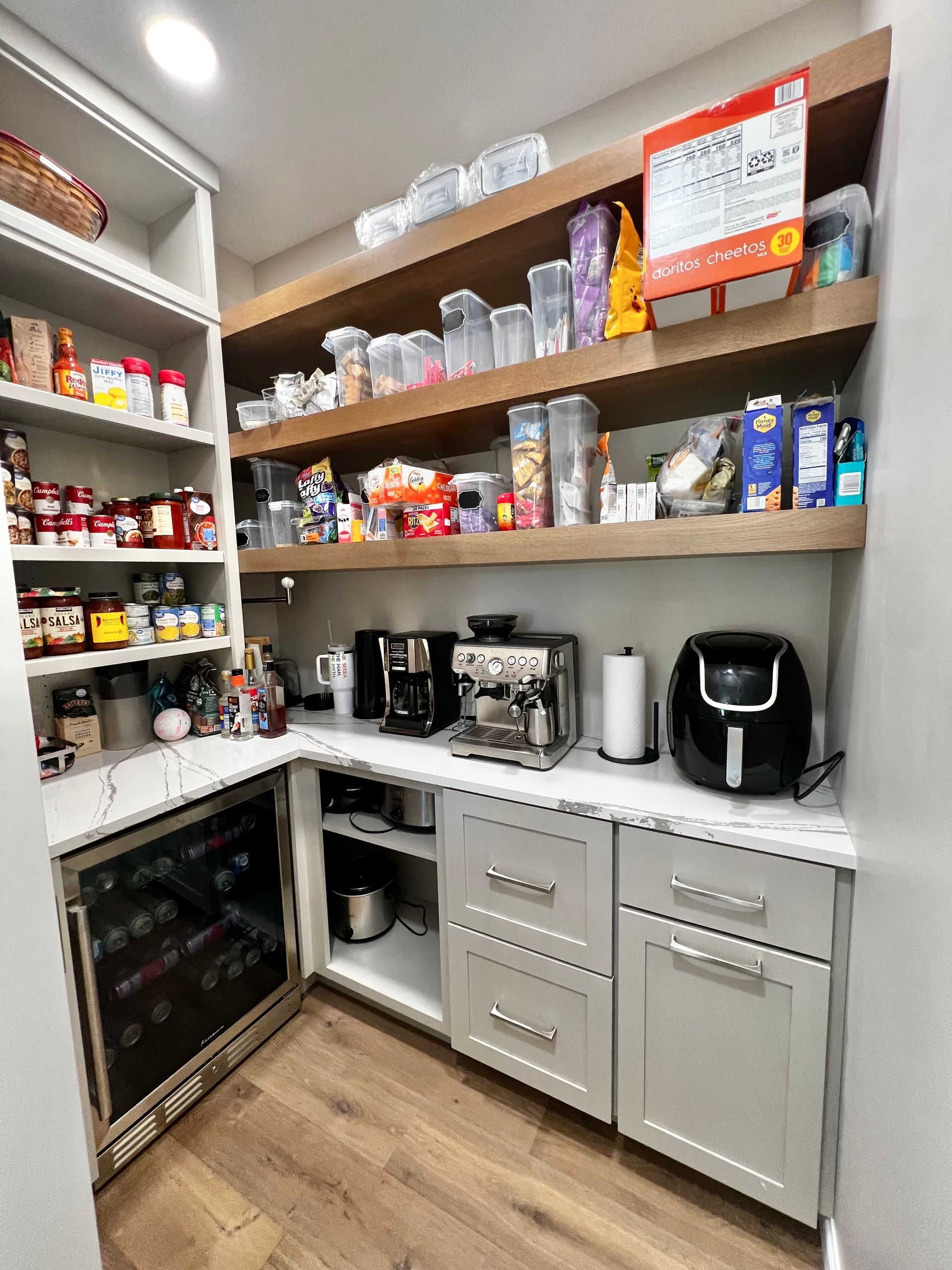 A organized kitchen pantry with open wooden shelves holding food containers and a counter with appliances and drawers.