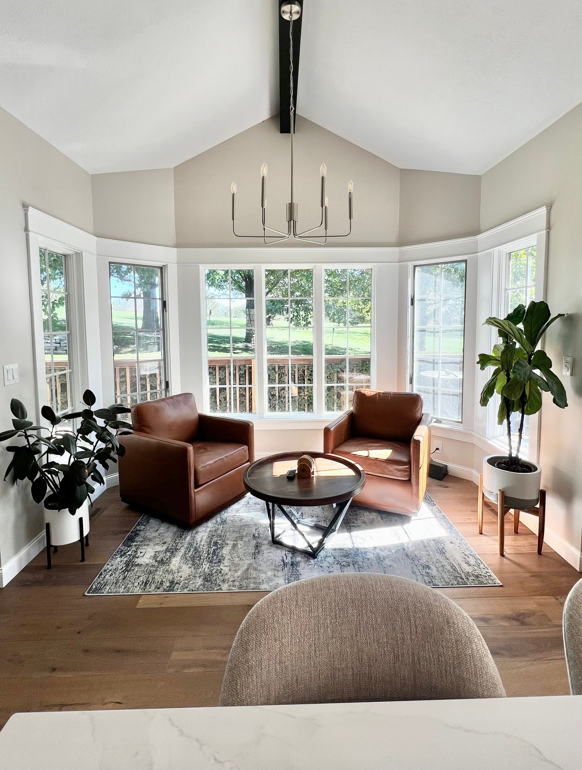 Sun-filled nook with two leather armchairs, a round coffee table on a patterned rug, and houseplants by bay windows.