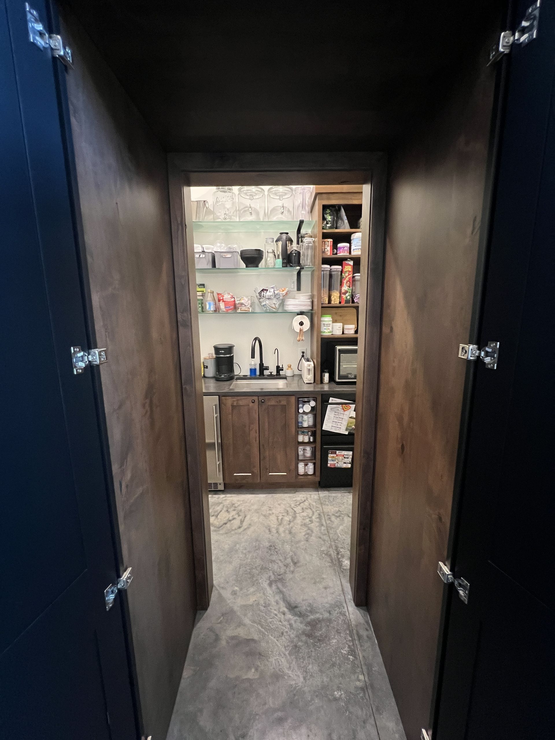 A doorway viewed from a dark passage reveals a brightly lit pantry or kitchenette with shelves, a counter, and cabinets.