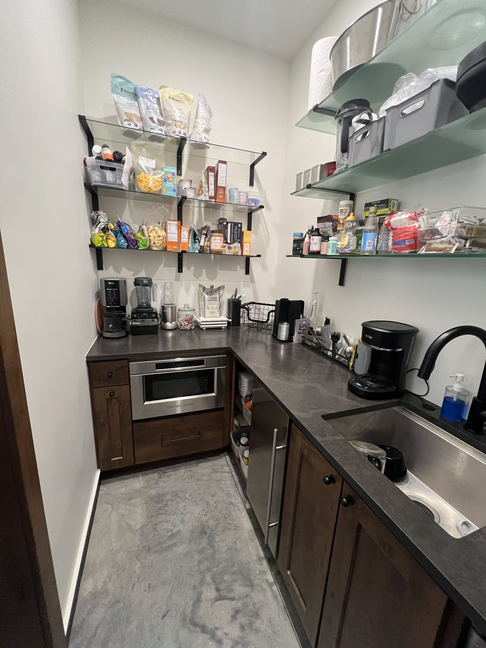 A galley-style kitchenette with dark wooden cabinets, a sink, a built-in microwave, and open shelves stocked with snacks.