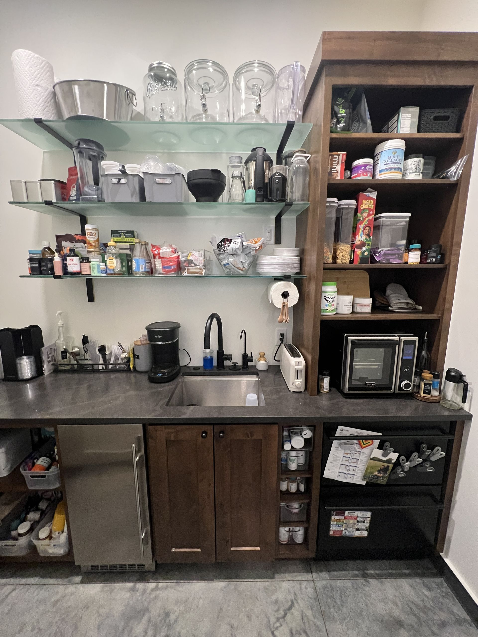 A kitchen break room featuring dark wood cabinets, open glass shelving, a sink, microwave, mini fridge, and snack storage.