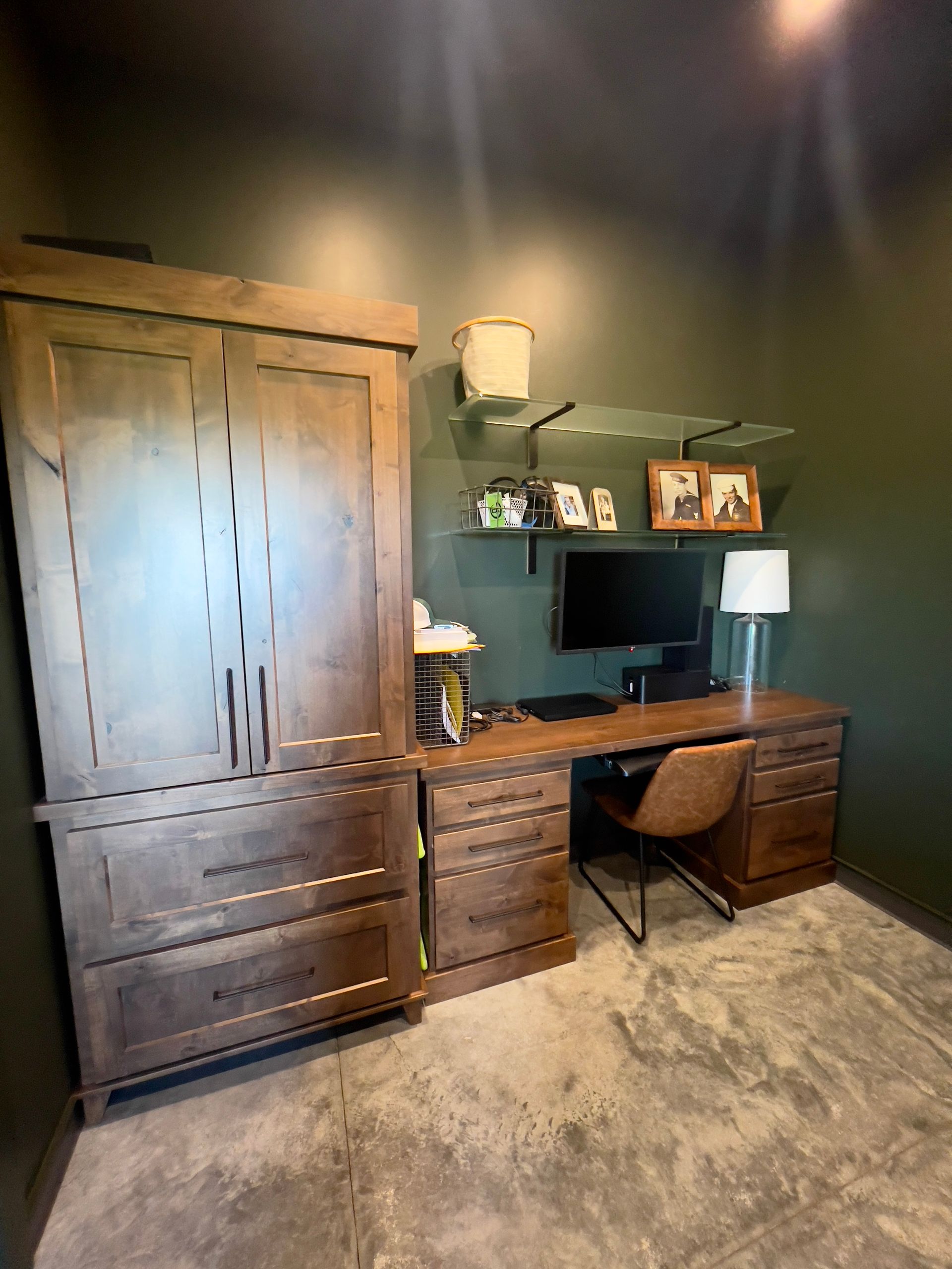 A home office with dark green walls, a large wooden wardrobe, a desk with a computer monitor, and a brown leather chair.