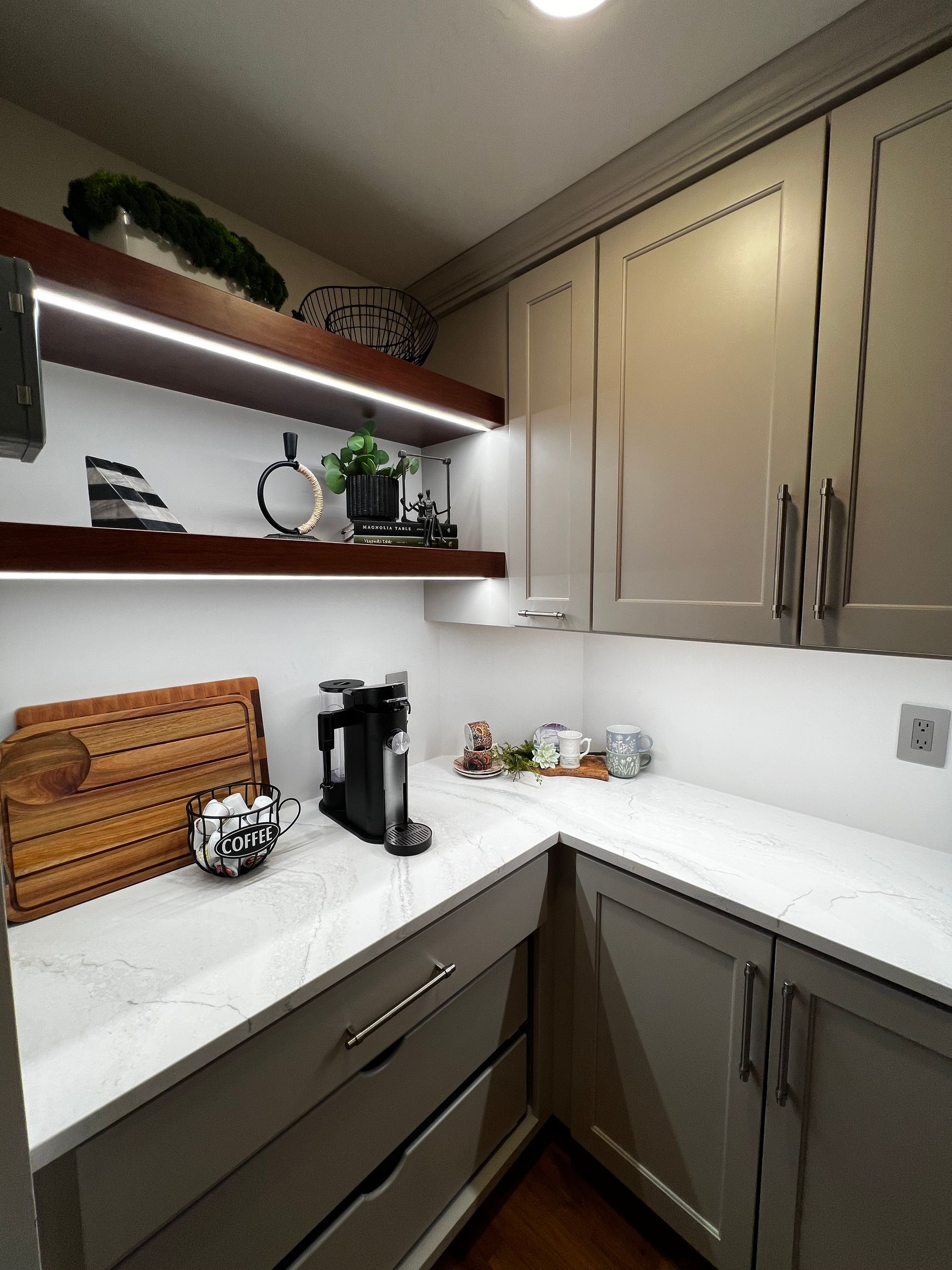A small, modern kitchen corner featuring gray cabinets, white marble countertops, and illuminated wood floating shelves.